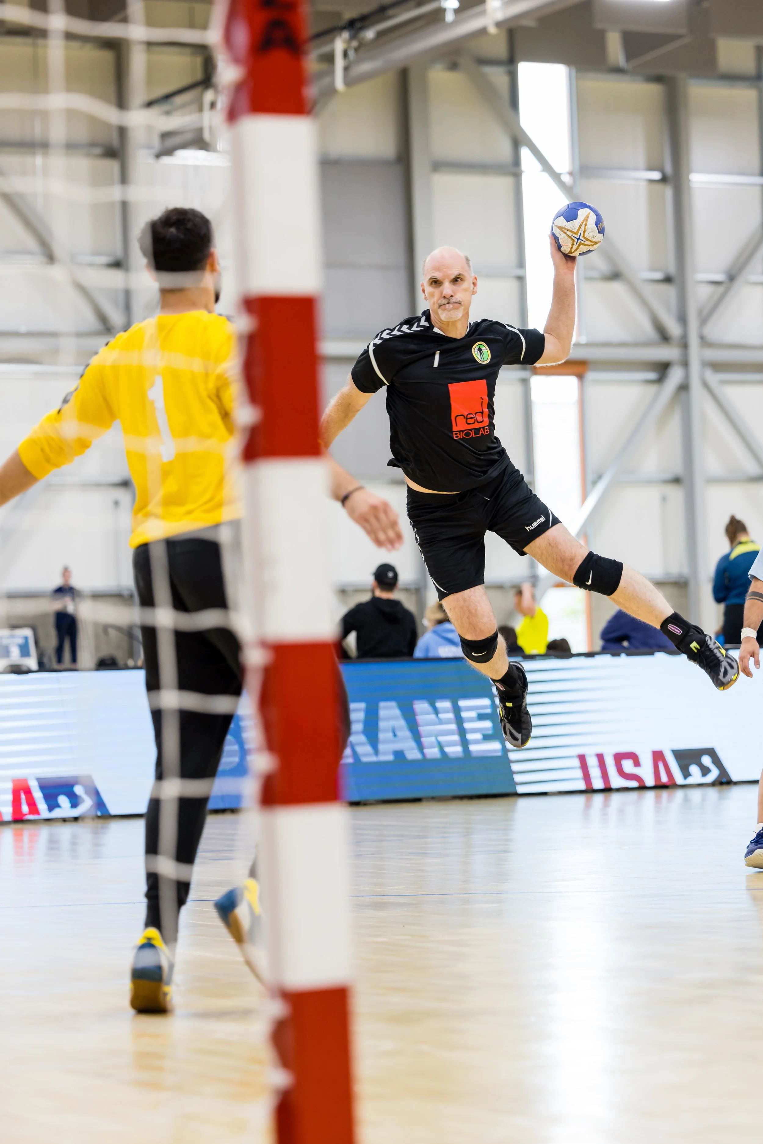 A man in a black sports uniform jumping to hit a volleyball during a game inside a gymnasium. A player in a yellow jersey is visible in the foreground, with a net and an indoor sports arena in the background.