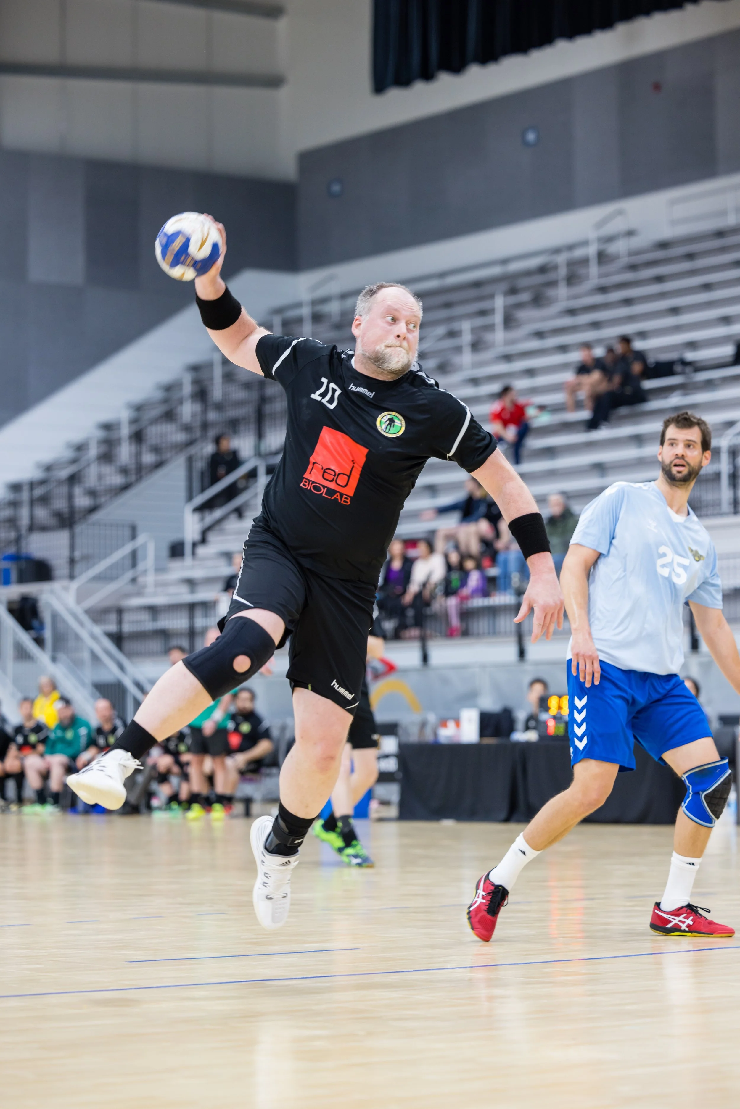 A man with a beard wearing a black sports uniform with the number 10 is playing indoor volleyball, jumping to hit the ball, with spectators in the background.