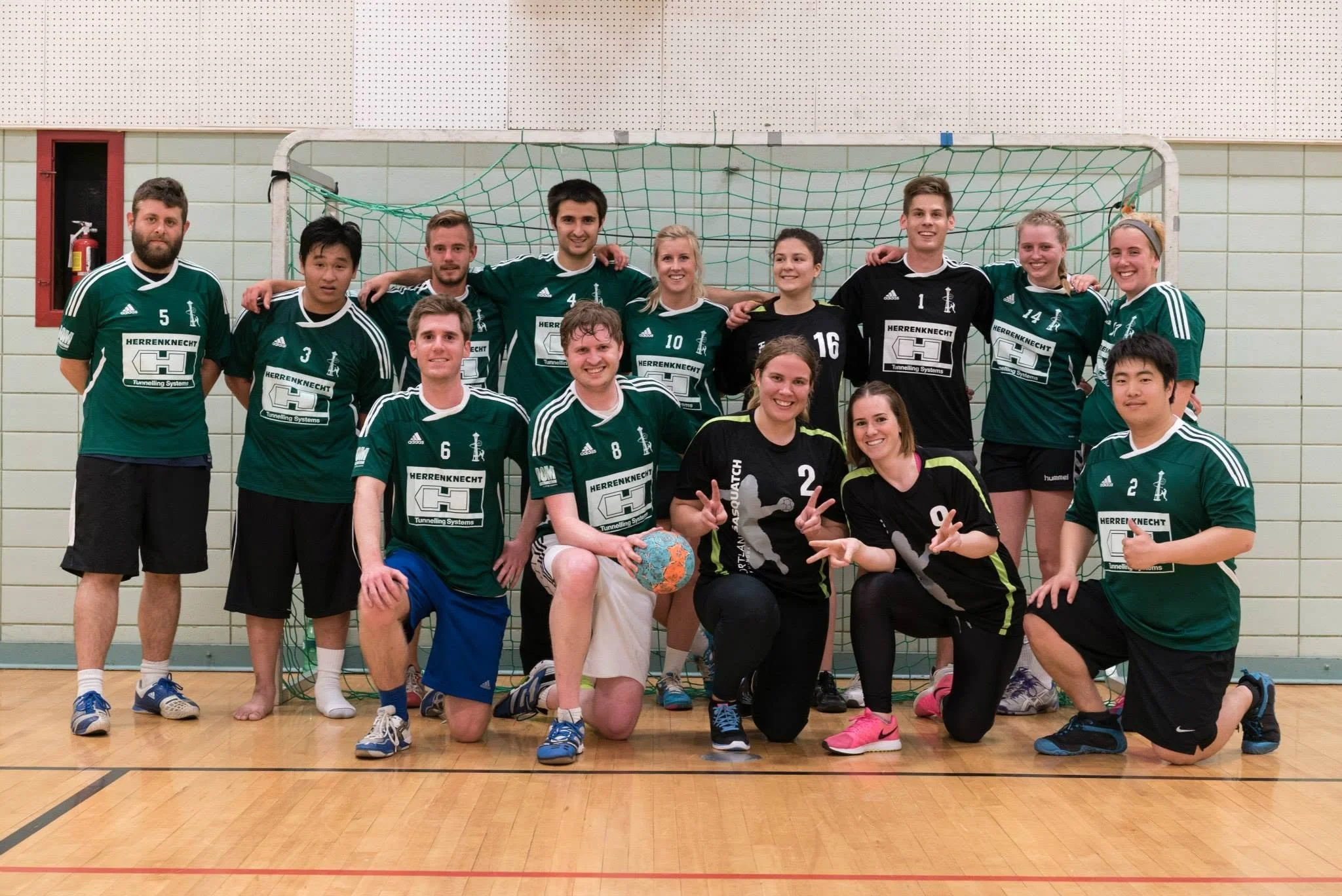 Group of twelve people, including men and women, posing for a photo in an indoor sports hall, some kneeling and others standing, in front of a handball goal, wearing sports uniforms.