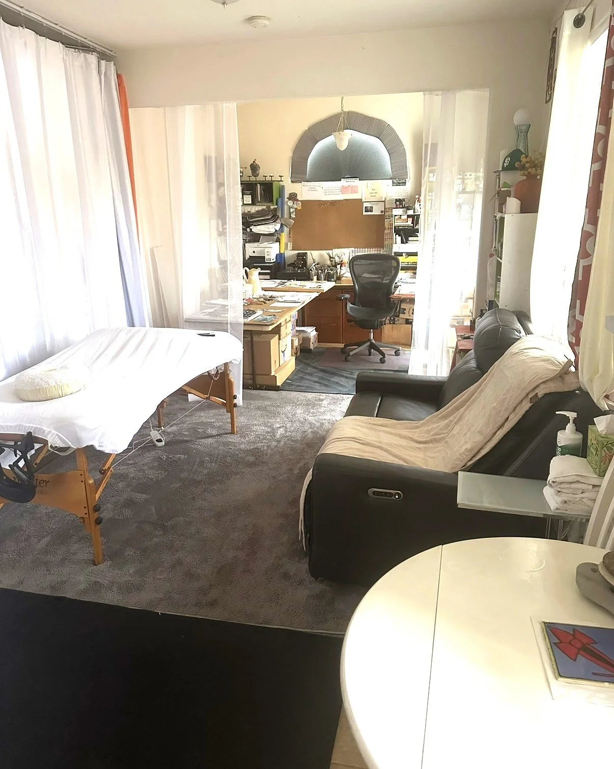 A small medical examination room with a massage table on the left, a black leather sofa with a beige blanket on the right, a white table with a book, and a workspace with a desk and office chair in the background.
