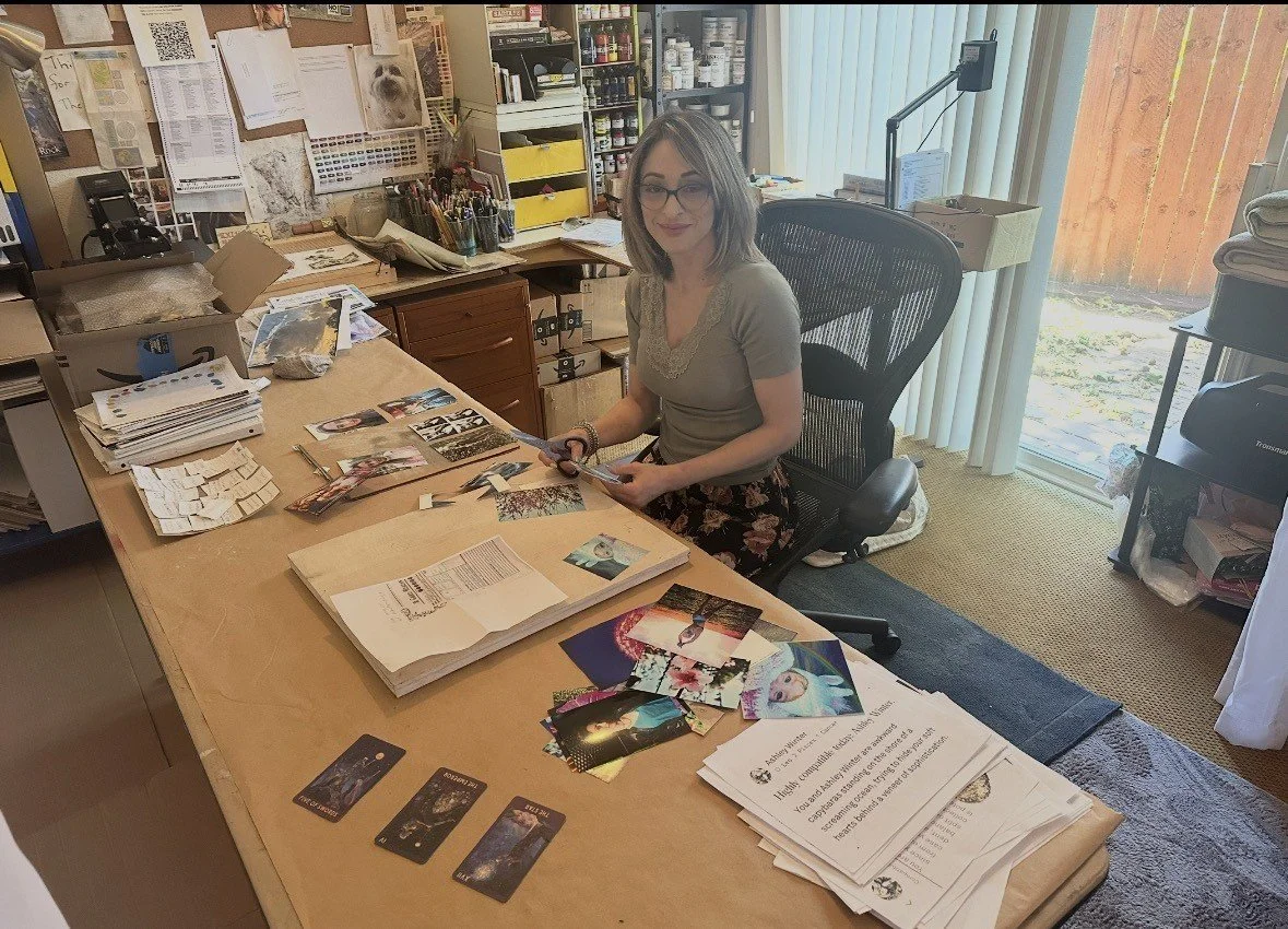 A woman is sitting at a cluttered desk in an office, cutting photographs with scissors. The desk has various papers, photographs, and office supplies on it. Behind her, there are shelves filled with boxes, bottles, and office materials. A large window with vertical blinds is to the right, letting in natural light.