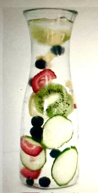 A glass pitcher filled with water and sliced fruits including strawberries, blueberries, kiwi, cucumber, lemon, and lime.