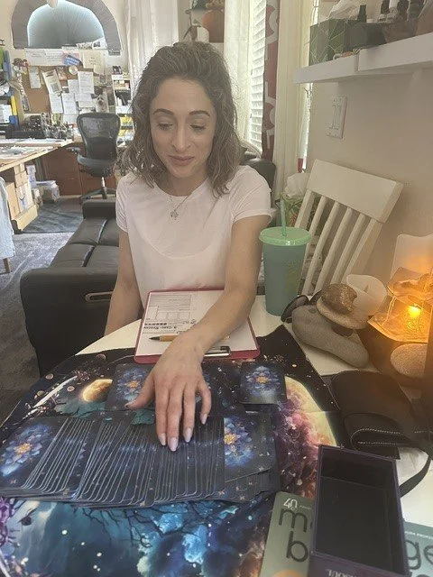A woman with curly hair wearing a white t-shirt sitting at a table in a room that appears to be a cafe or office. She is reaching out with her left hand to touch a collection of tarot cards laid out on a galaxy-themed mat. The table has a green reusable cup with a straw, some stones, a small lamp, and a smartphone. The background shows shelves and bulletin boards with various papers and items.