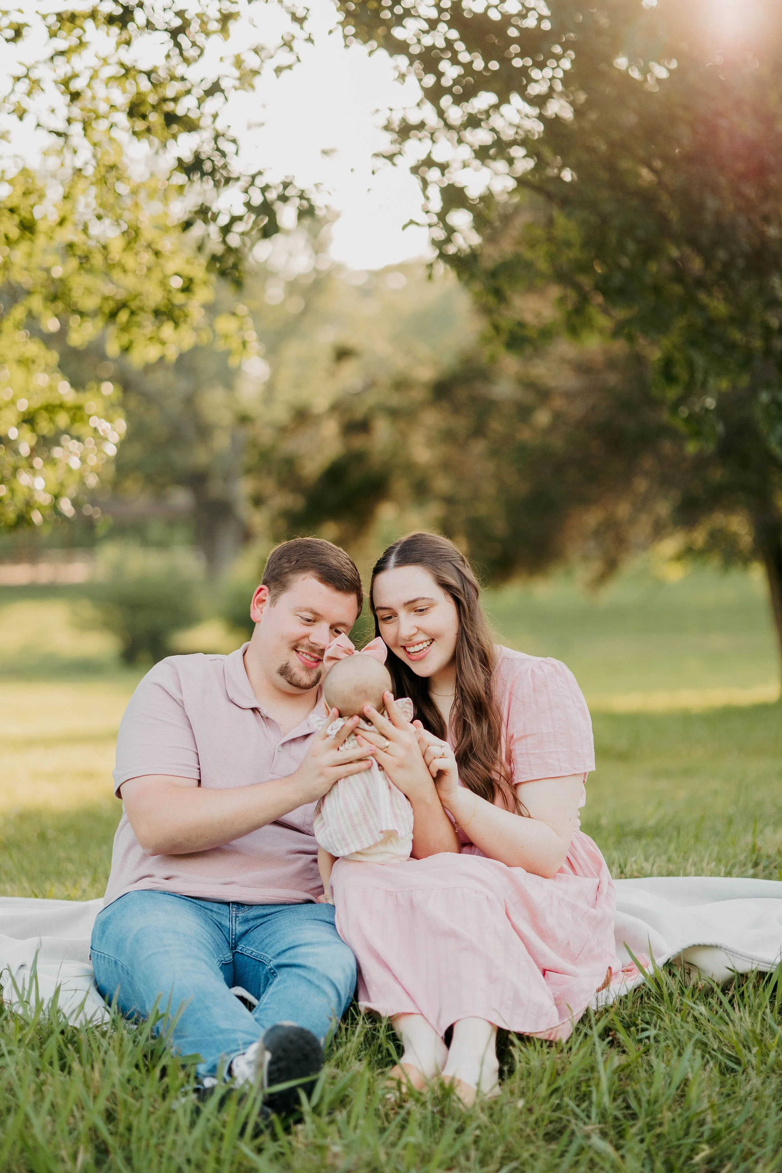 A family of three sitting on a white blanket outdoors in a grassy park during sunset, looking at their baby.