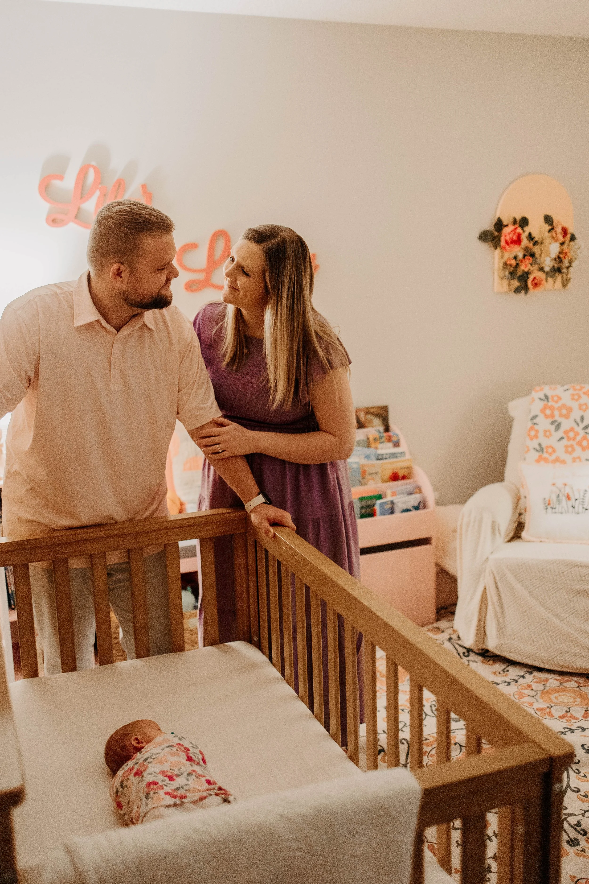 A man and woman standing by a crib, smiling and looking at each other in a nursery, with a baby sleeping in a crib in the foreground.