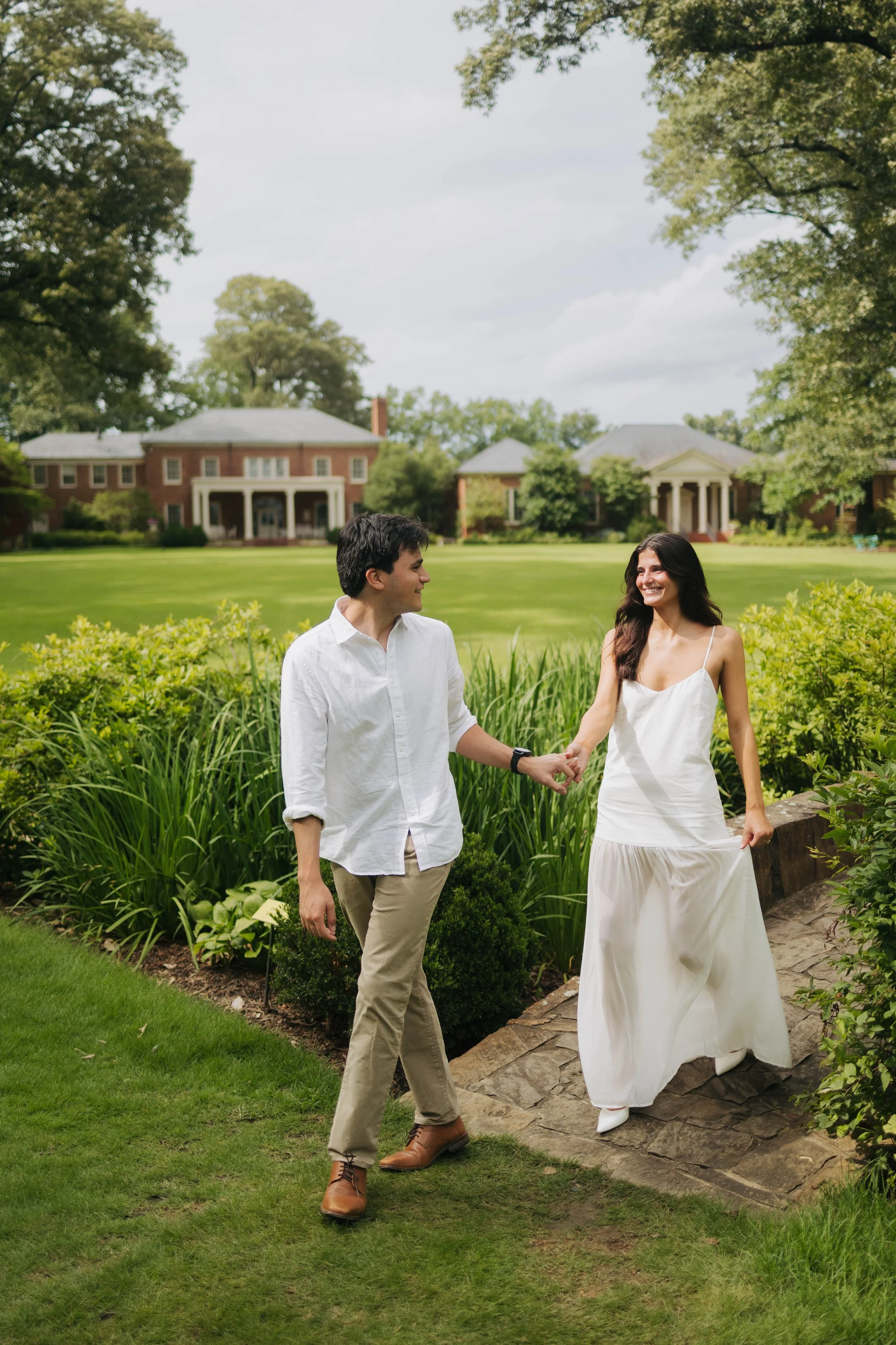 A couple standing outdoors on a garden path, holding hands and smiling at each other, with large green trees, a grassy field, and two brick houses with columns in the background.
