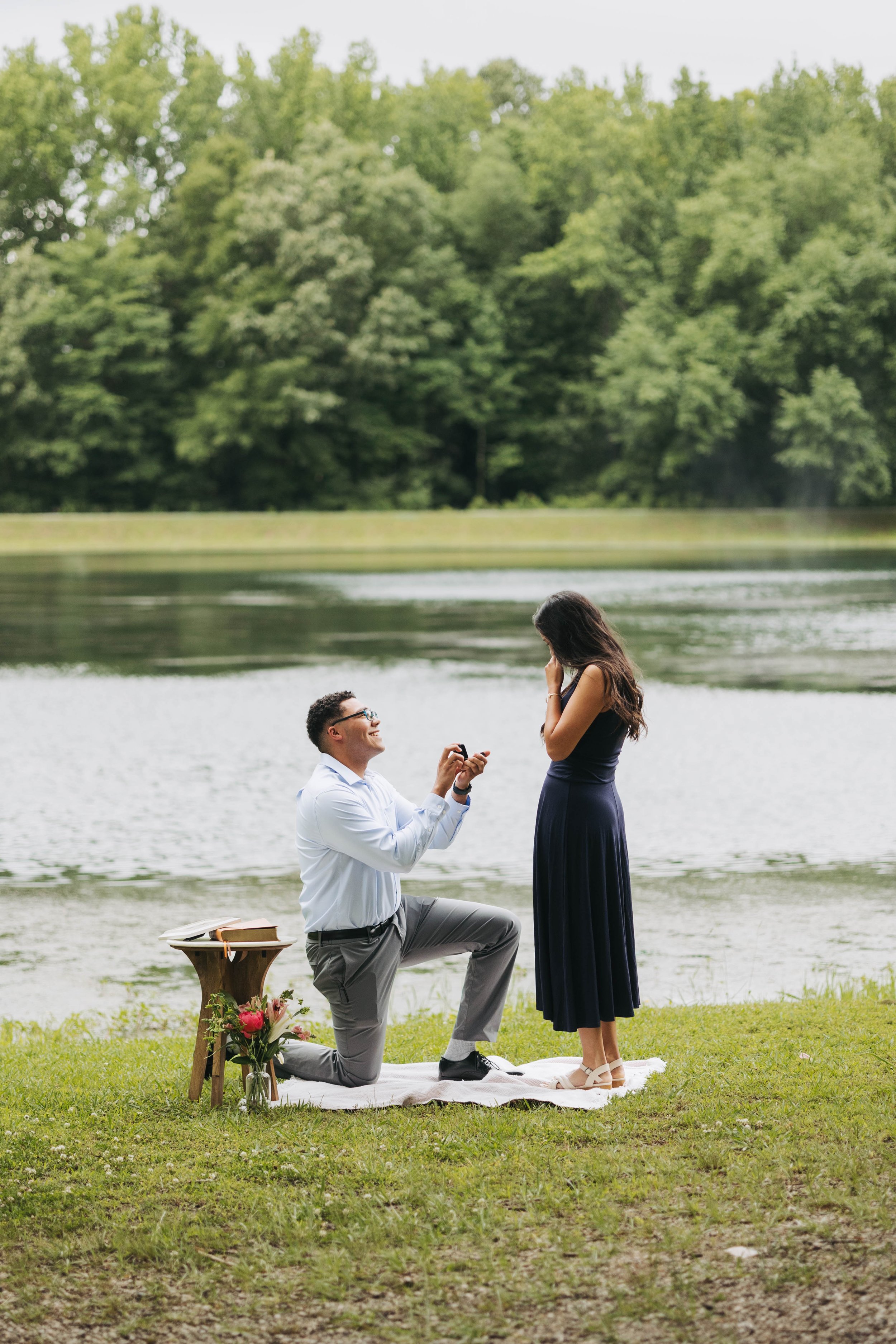 A man proposing to a woman by a lake during the daytime. The man is kneeling on one knee, holding a ring, while the woman stands before him, covering her mouth in surprise. There is a small table with a bouquet of flowers and books beside them, on a 