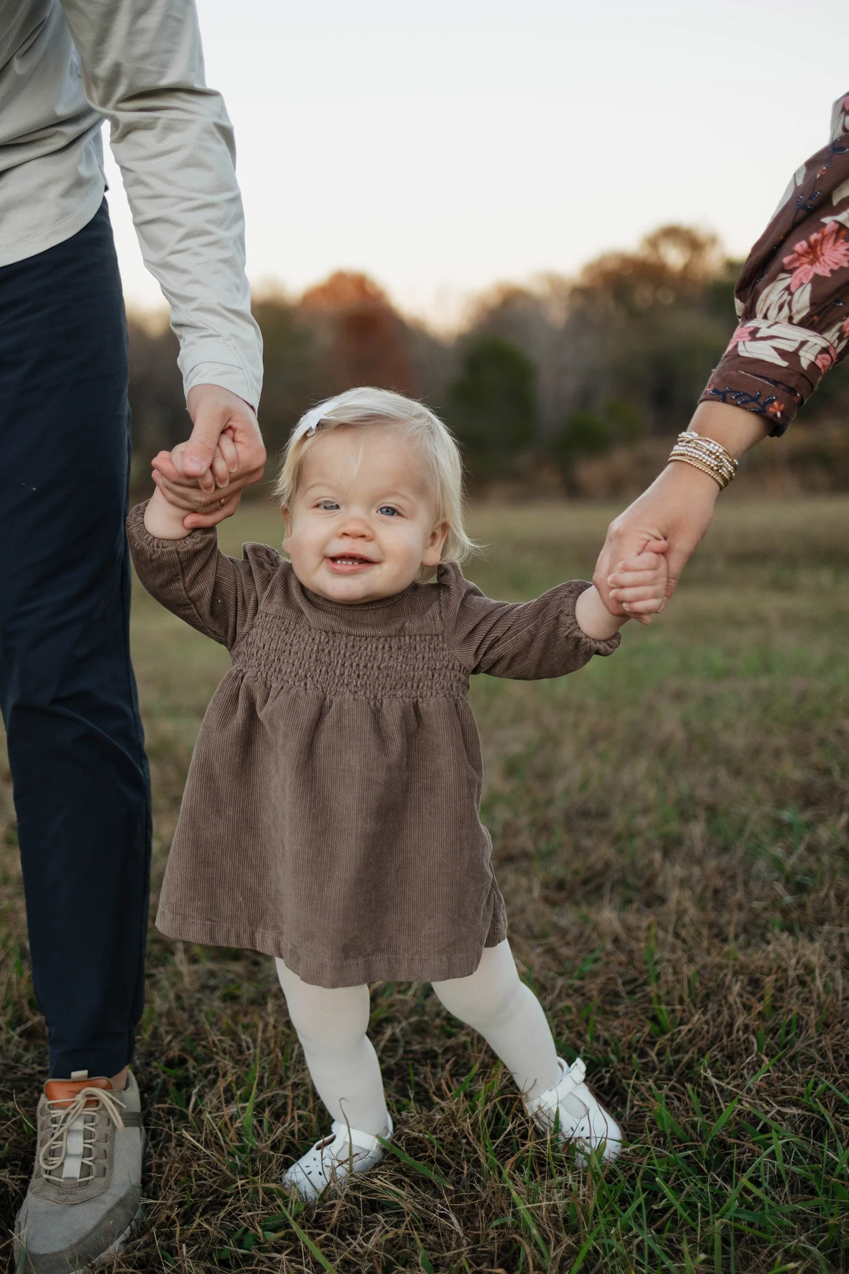 A young girl standing outdoors on grass, holding both adults' hands, with the sun setting behind trees in the background. The girl is smiling and wearing a brown dress, white tights, and sneakers.