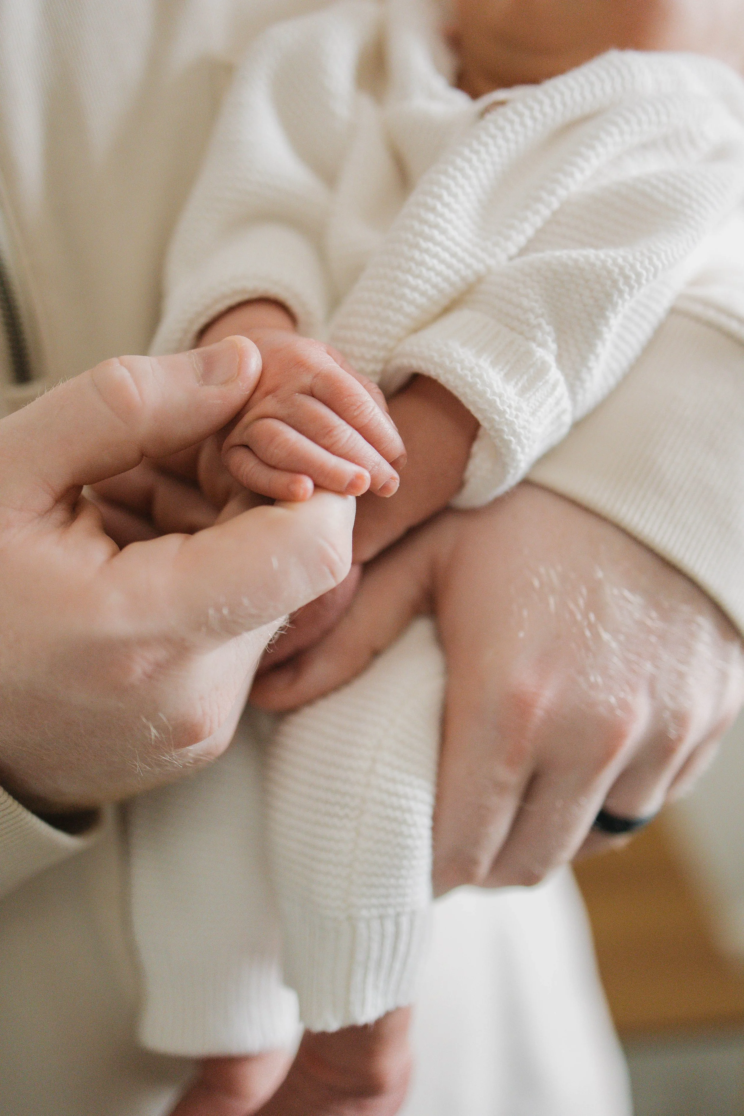 Close-up of an adult hand holding a baby's hand, with the baby wearing a white knitted sweater.