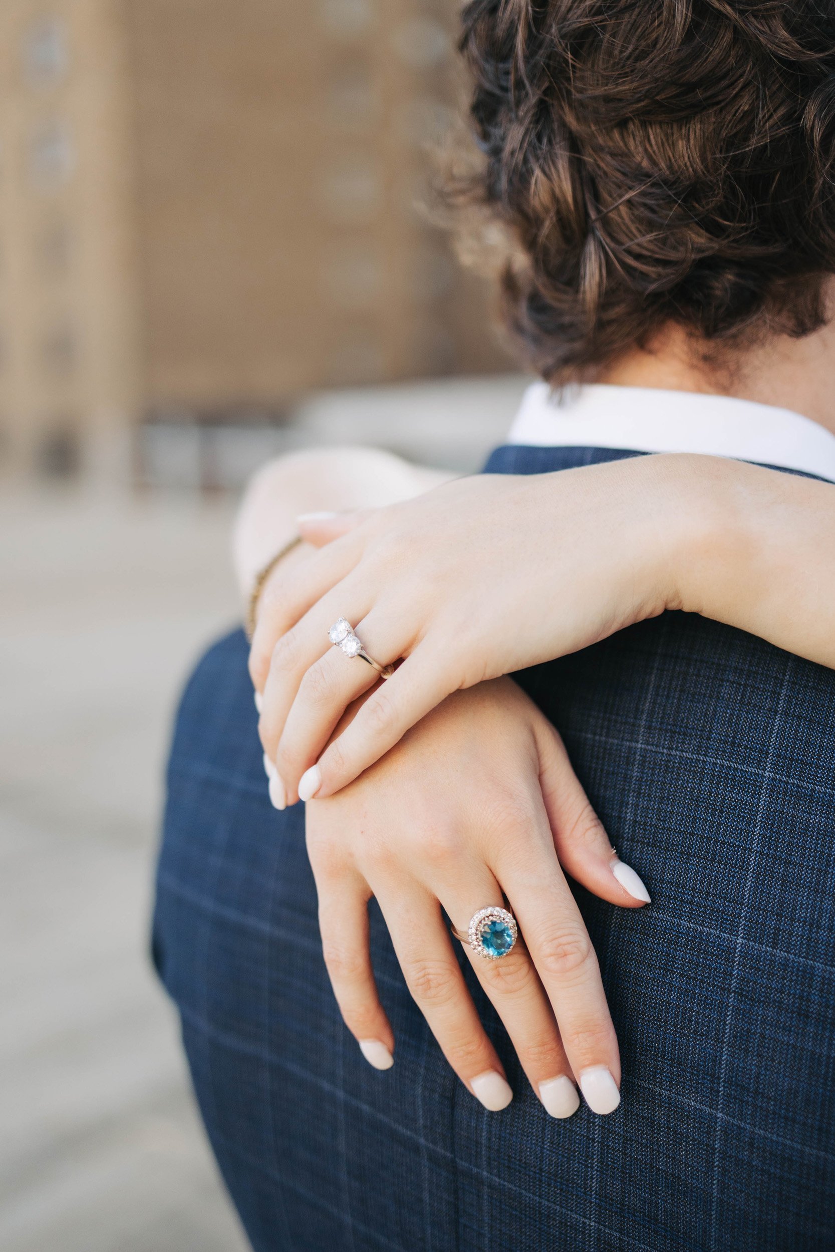 Close-up of a person's hands with rings, resting on their shoulder, in a formal outfit. The person has short, curly brown hair, and the background is blurred.