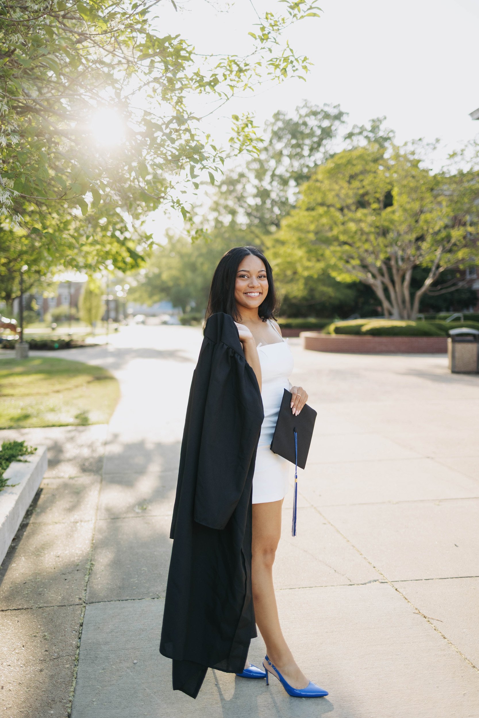A young woman in a white dress and blue high heels stands outside on a sidewalk, holding a black graduation cap and draped in a black graduation gown, smiling. There are trees and a park-like setting in the background with the sun shining through lea
