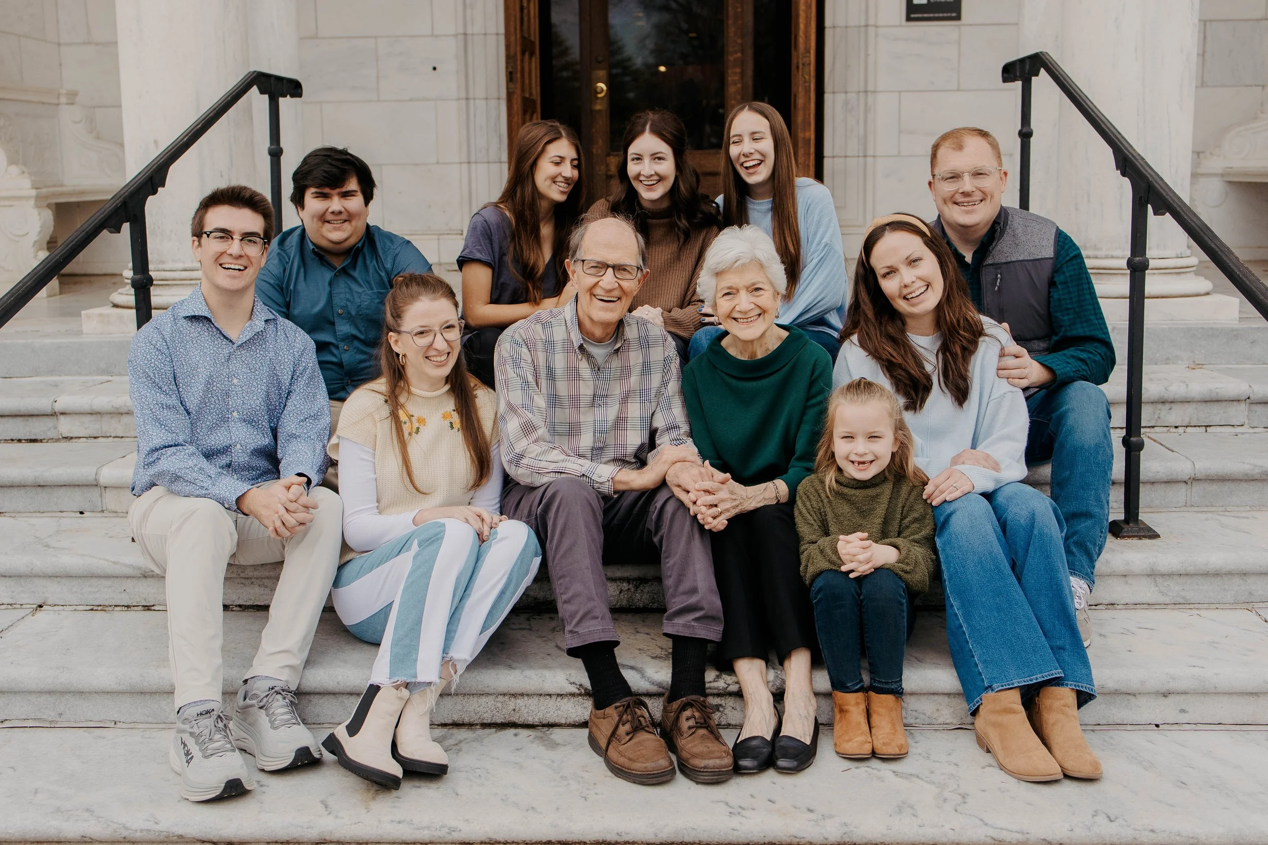 Group of twelve people, including children, teenagers, and seniors, sitting on stone steps in front of a building with large wooden entrance doors, smiling and posing for a photo.