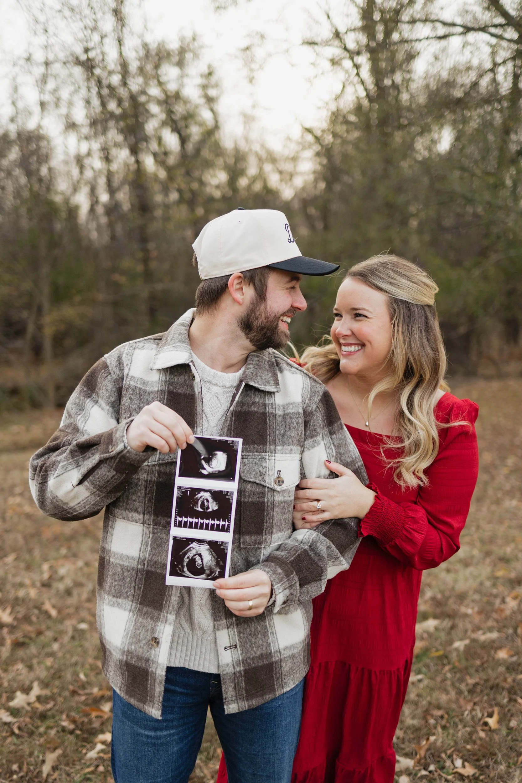 A smiling couple standing outdoors, with the woman in a red dress and the man holding ultrasound images. The woman is embracing the man, and they are looking at each other with happiness, with trees and autumn leaves in the background.