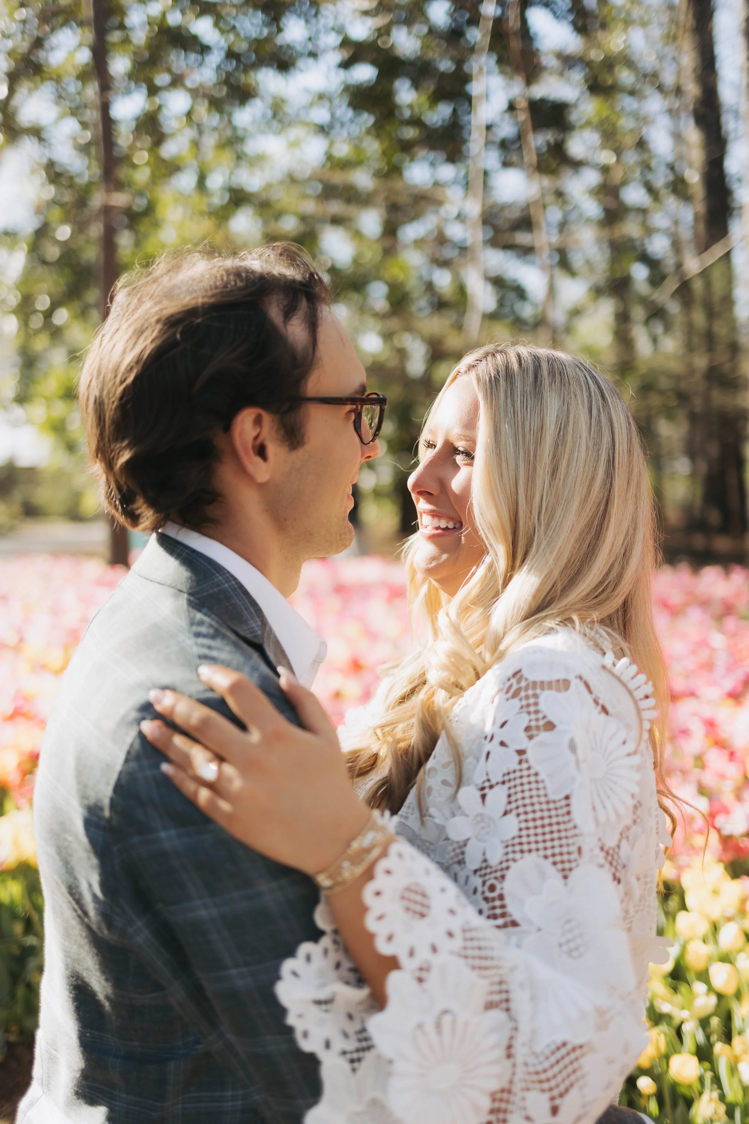 A couple is embracing outdoors in a park, facing each other with smiles, surrounded by trees and flower beds.