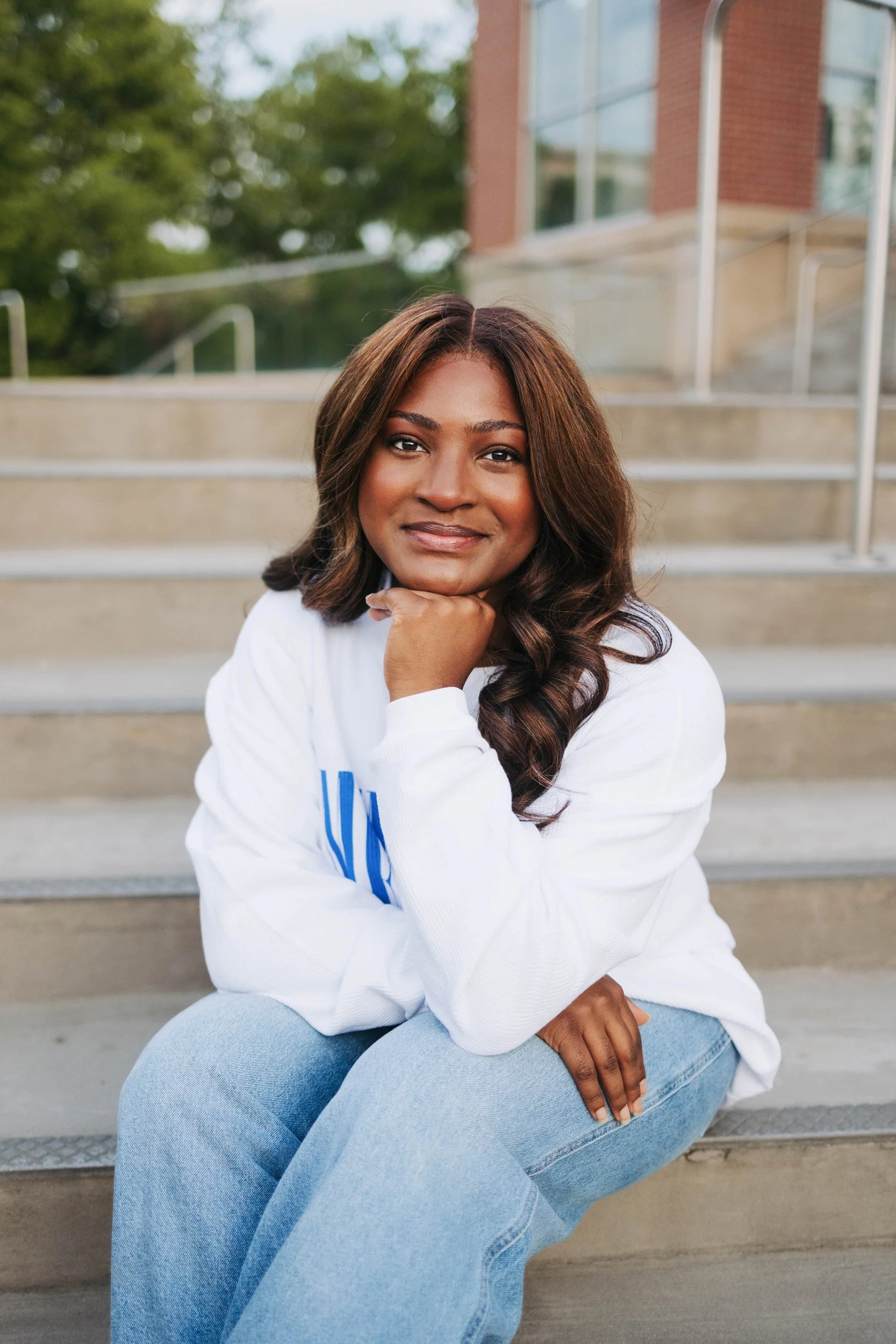 A woman with medium-length brown hair and a friendly smile sitting on concrete stairs outdoors, wearing a white sweatshirt and light blue jeans, with green trees and a brick building in the background.