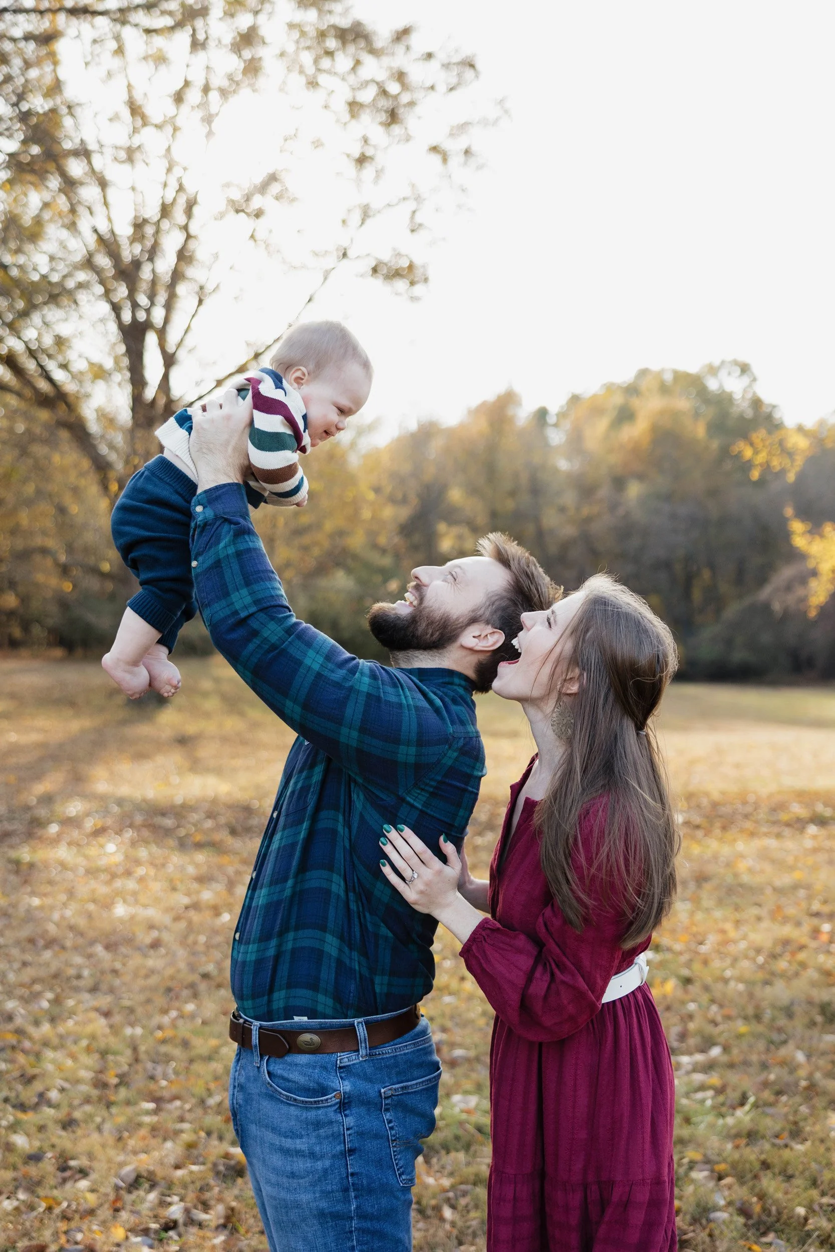 A man with a beard and a woman with long hair are standing outdoors in a park during fall. The man is lifting a baby boy into the air, and everyone is smiling and looking at each other.
