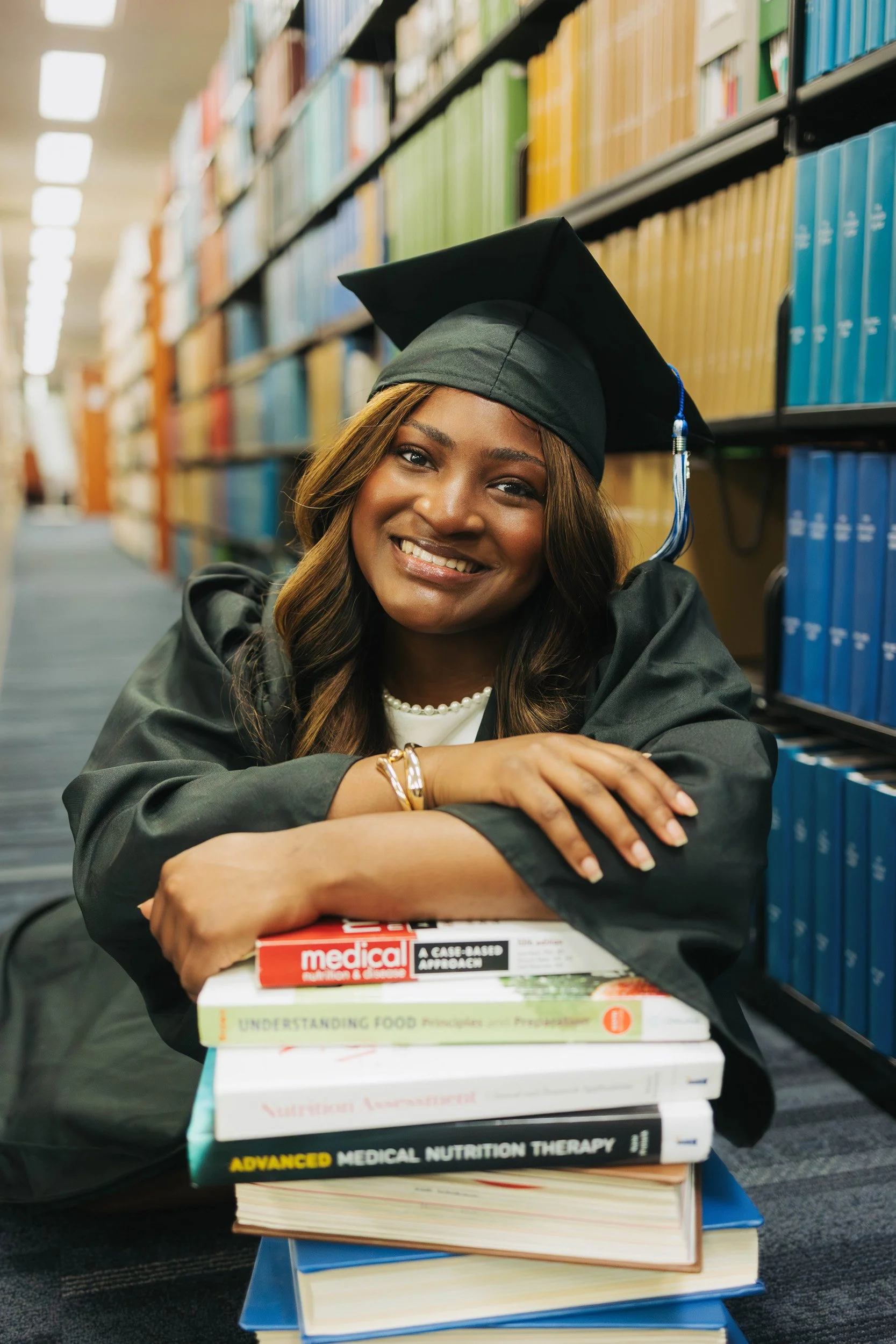 A smiling woman in graduation cap and gown sitting at a library with a stack of textbooks.
