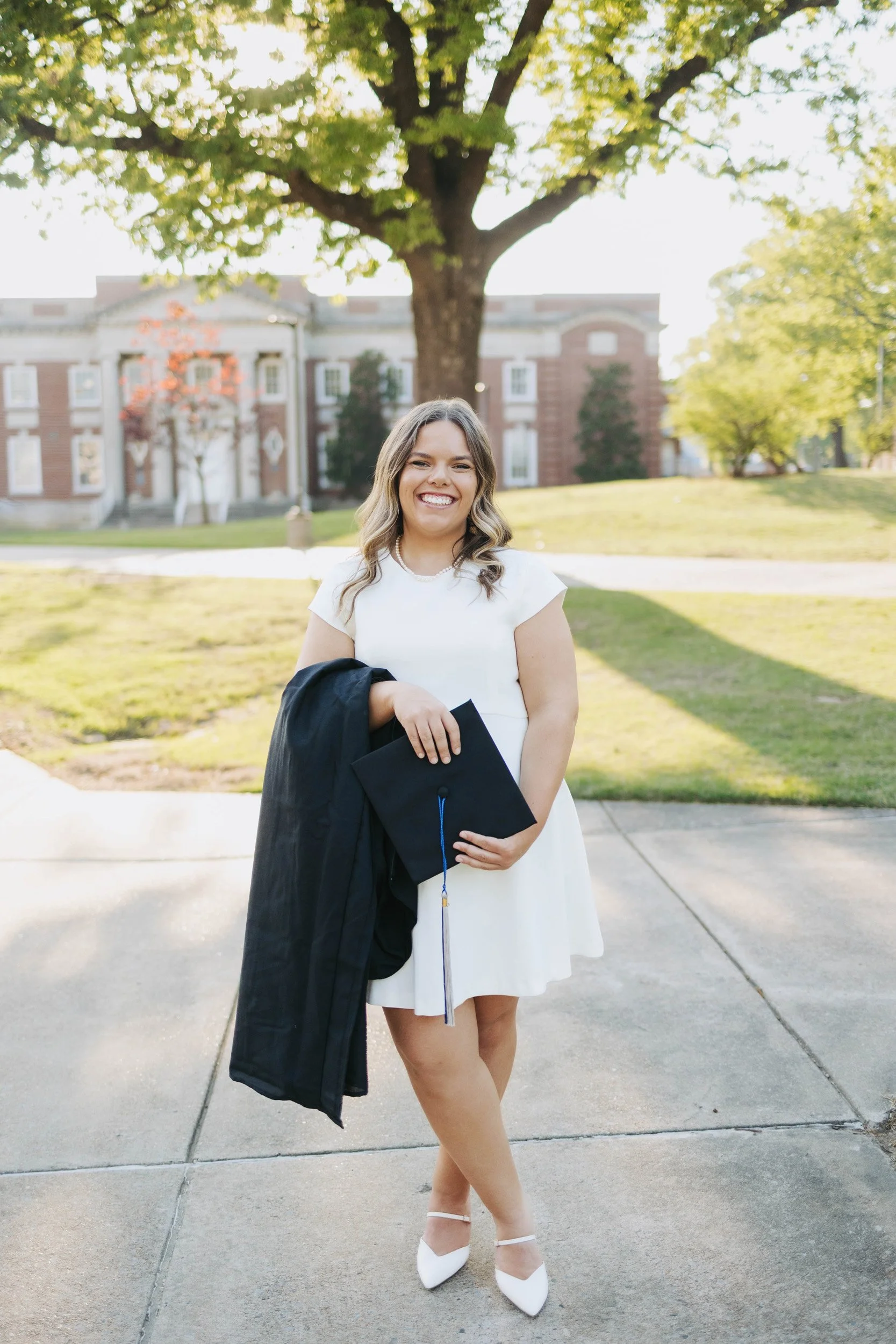 Young woman in white dress holding graduation cap and gown, smiling, standing outdoors in front of a large tree and a historic building, during daytime.