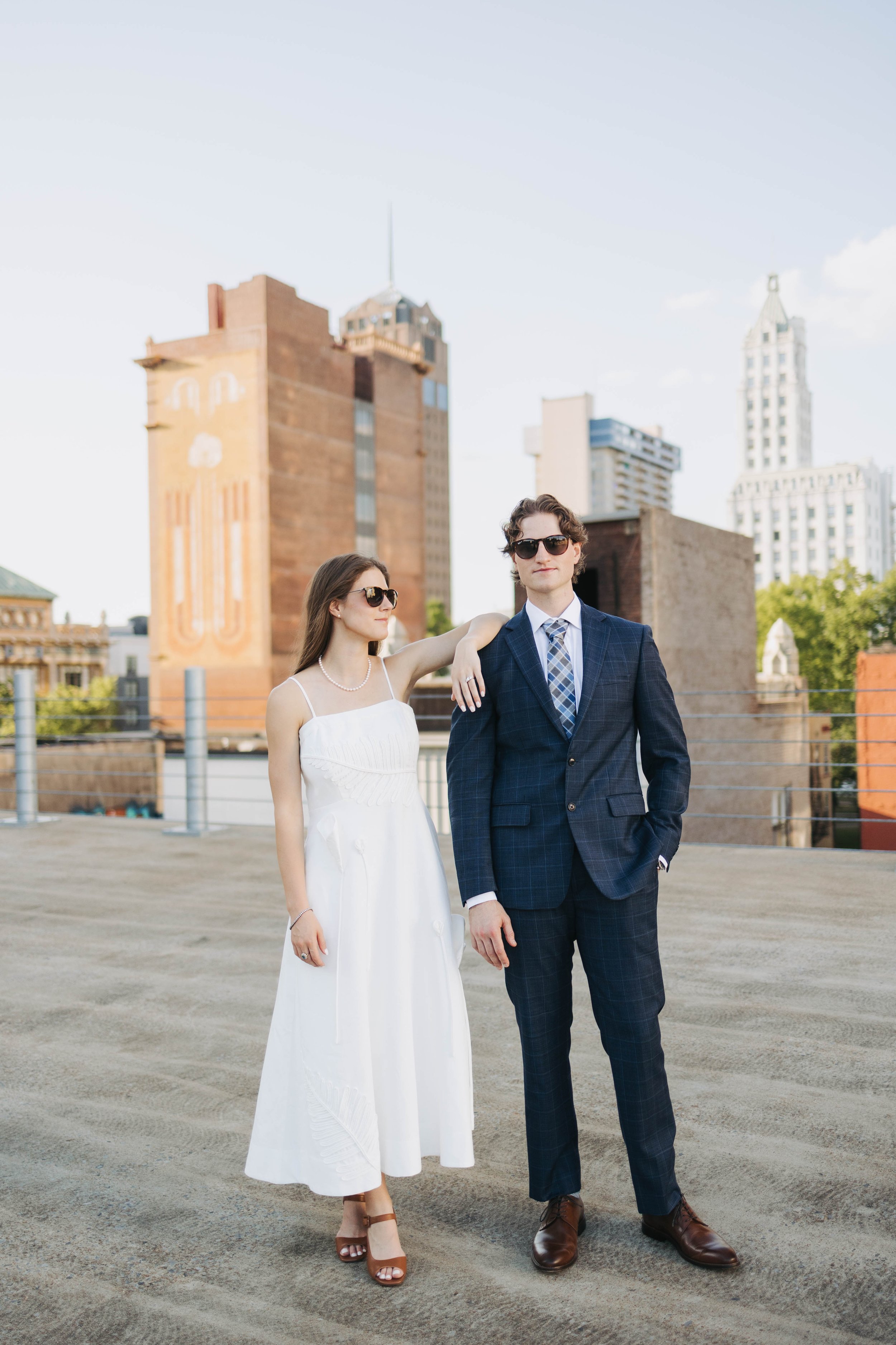 A man and woman dressed formally standing outdoors on a rooftop with city skyscrapers in the background. The woman wears a white dress and sunglasses, and the man wears a blue plaid suit, tie, and sunglasses. The woman has her arm around the man's sh