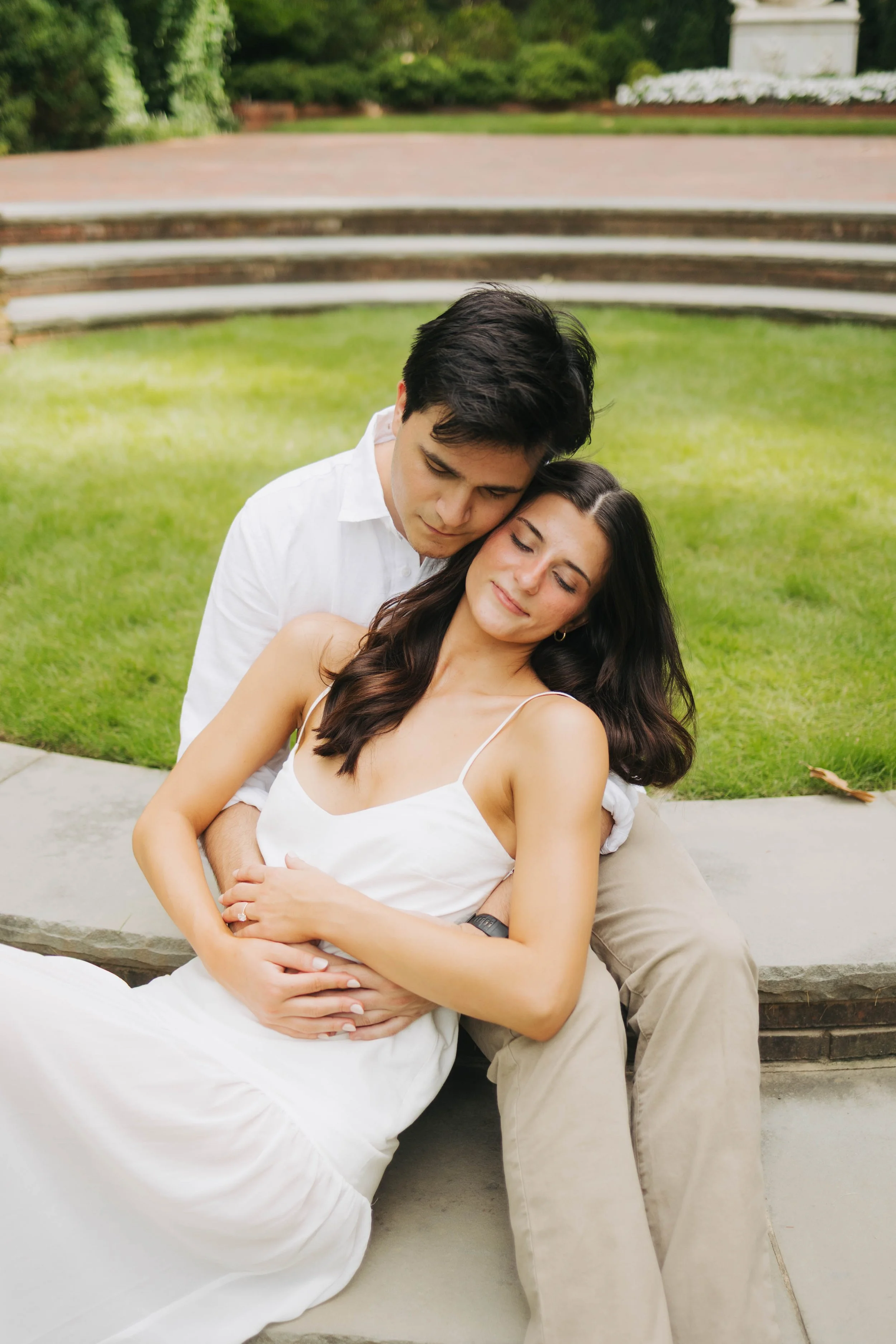 A young man and woman sitting outdoors on a stone bench, embracing and appearing relaxed and happy in a lush garden setting.