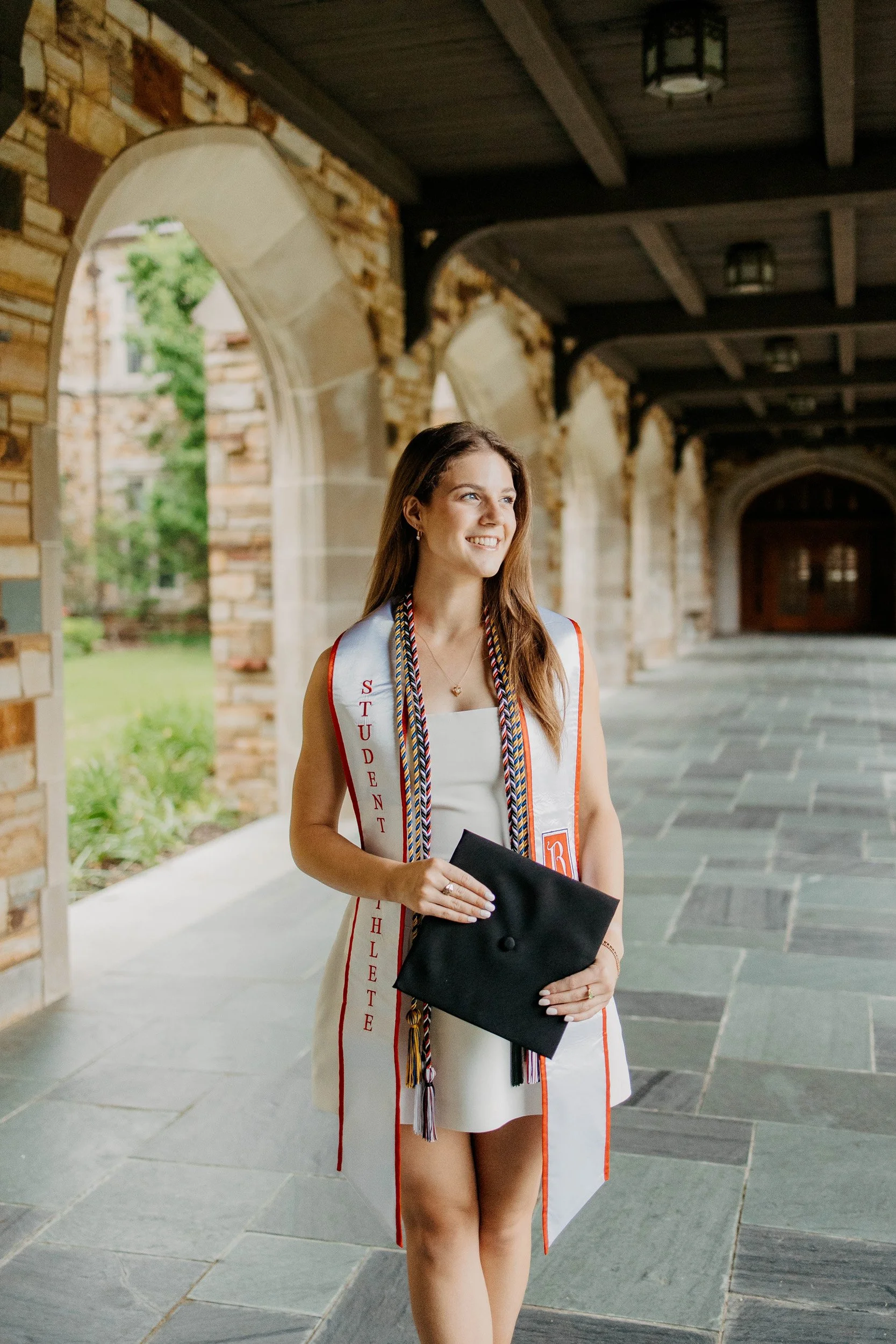 A young woman in a graduation cap and gown holding a diploma, standing under a stone archway in an outdoor setting with stone buildings and greenery in the background.