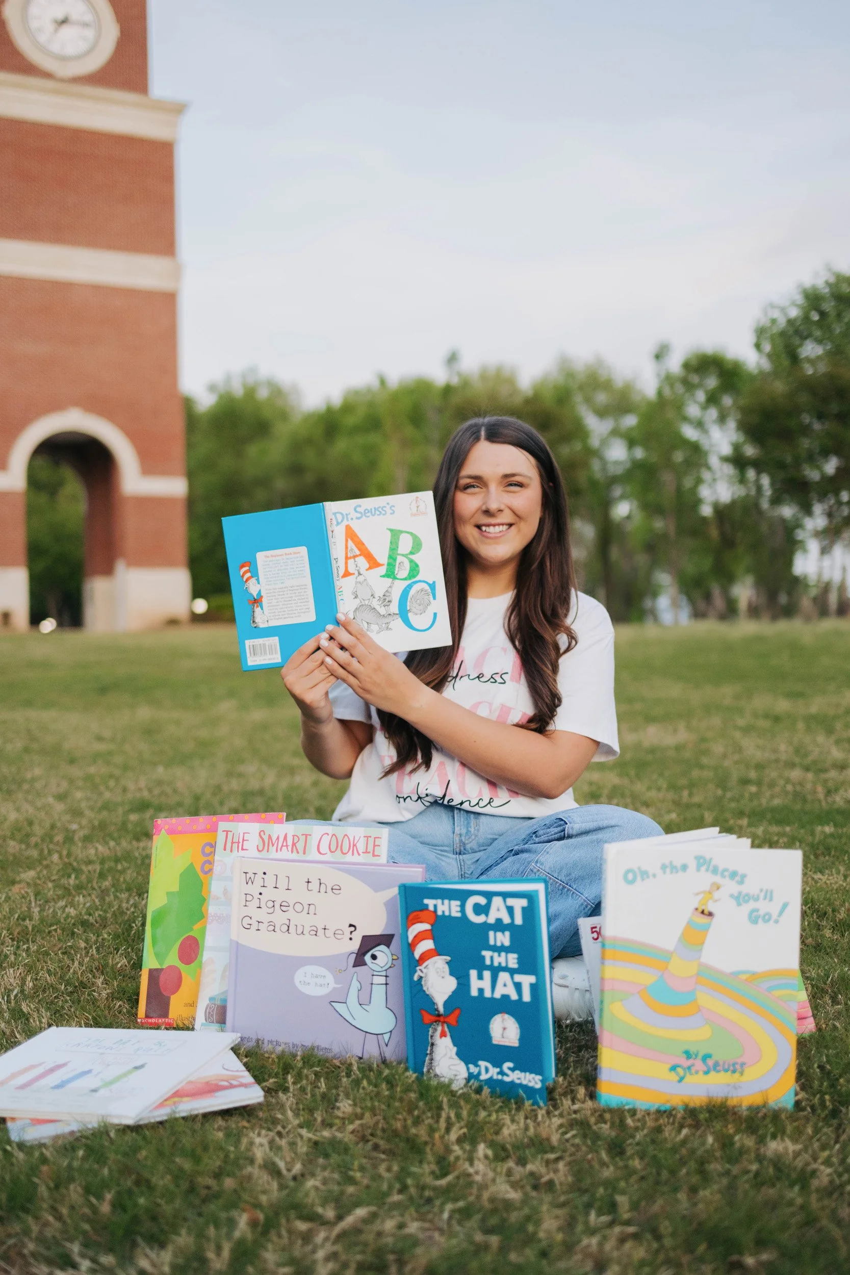 A young woman with long dark hair sitting on grass outdoors, holding a children's book and smiling at the camera. There are several children's books spread out on the grass in front of her, with titles such as "The Cat in the Hat" and "Oh, the Places