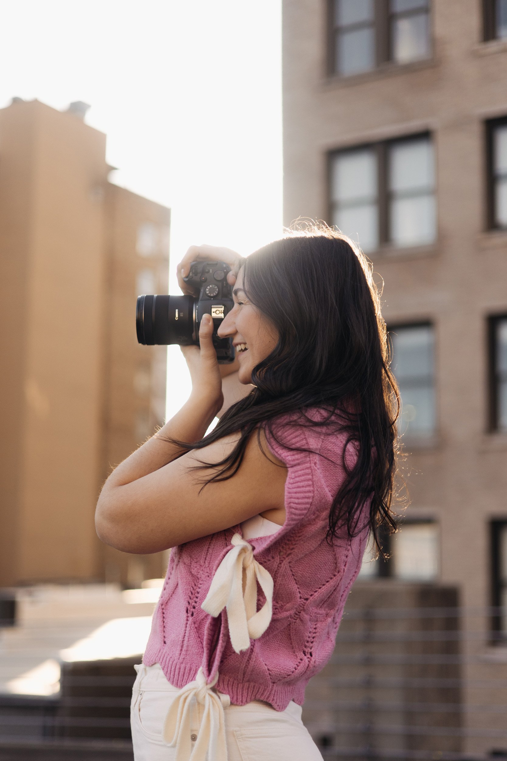 A woman with long dark hair is smiling and taking a photo with a camera during sunset. She is wearing a pink sleeveless knitted sweater with a ribbon on the side and white shorts. The background features multi-story buildings with windows and a set of stairs.