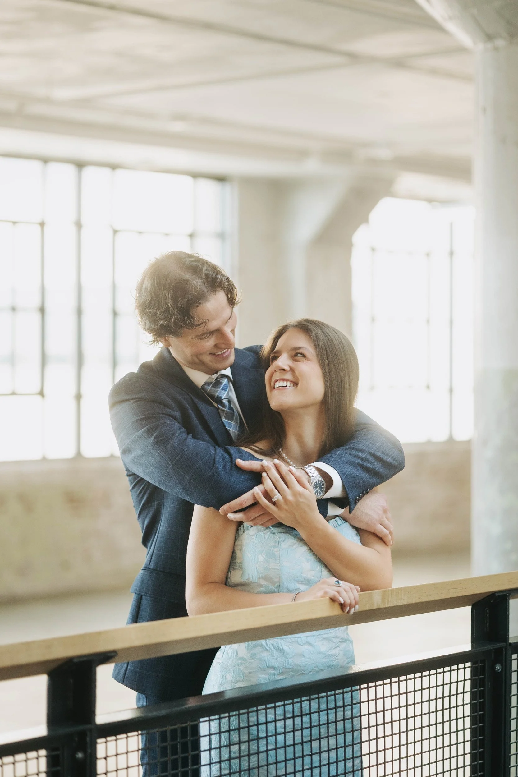 A man and woman smiling and embracing each other inside a bright, spacious building with large windows.
