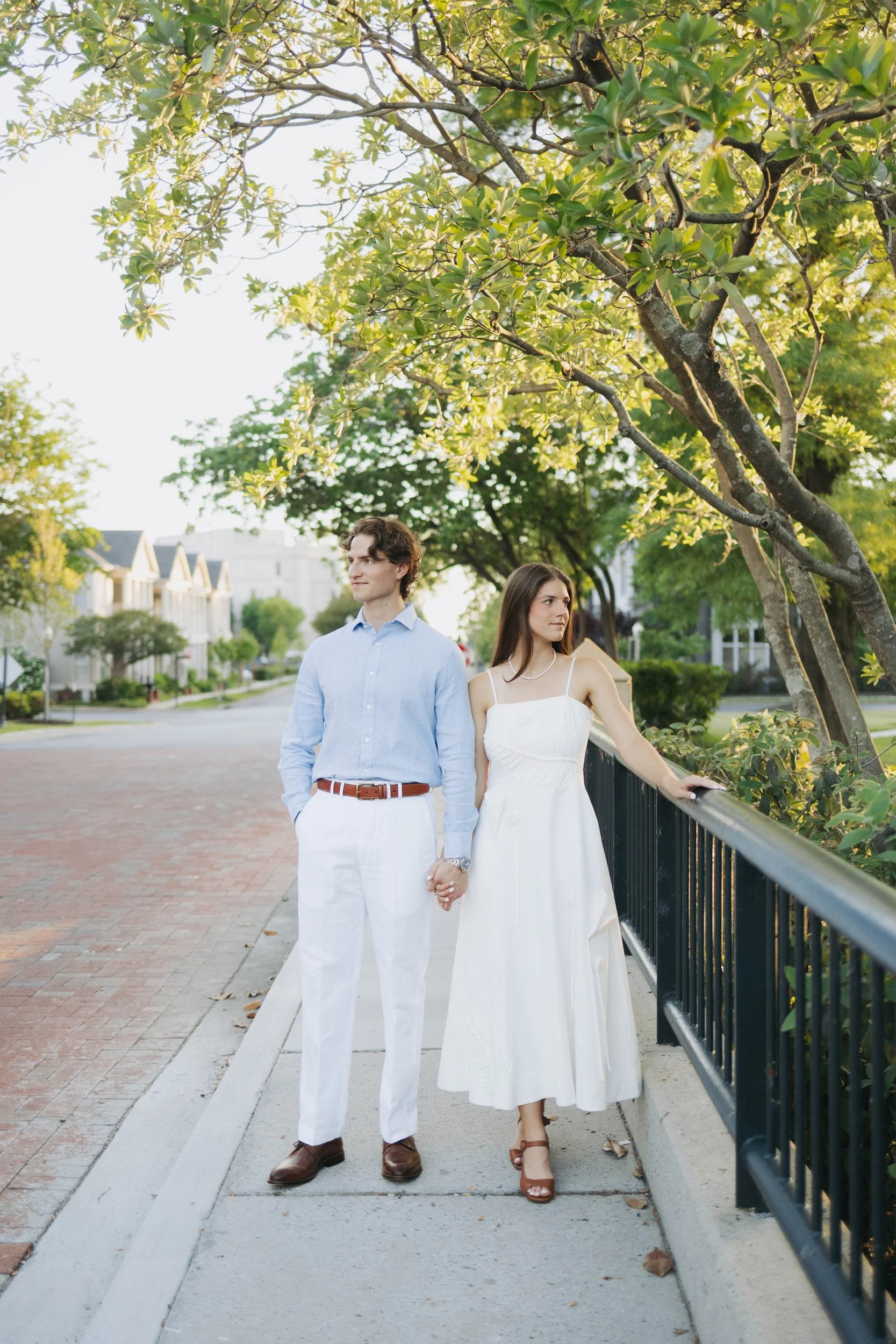 A young man and woman holding hands and standing on a sidewalk beside a black metal railing, with trees and suburban houses in the background, during daylight hours.