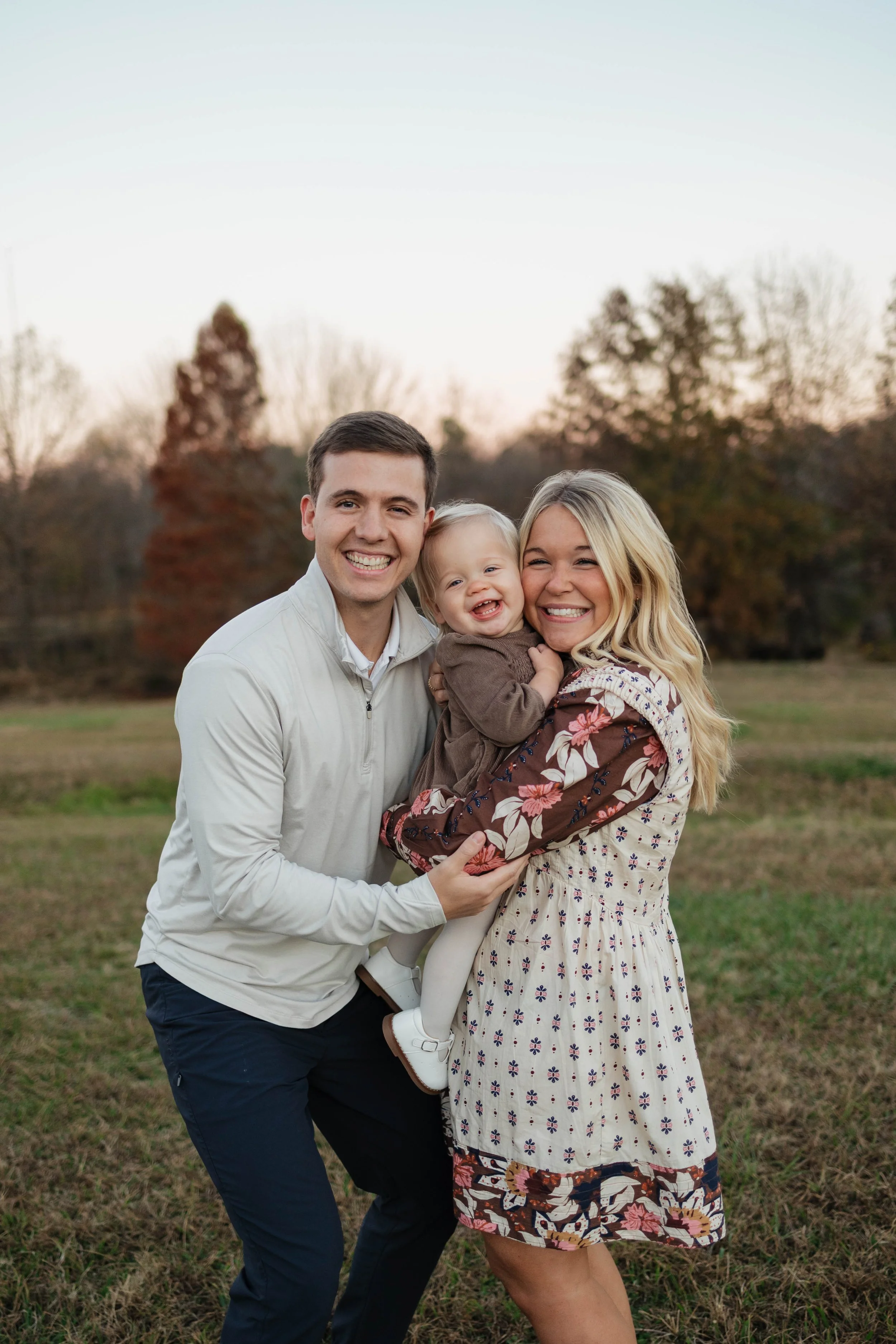 A happy family of three posing outdoors during sunset in a park, with trees and a grassy field in the background. The family includes a man, woman, and a young girl.