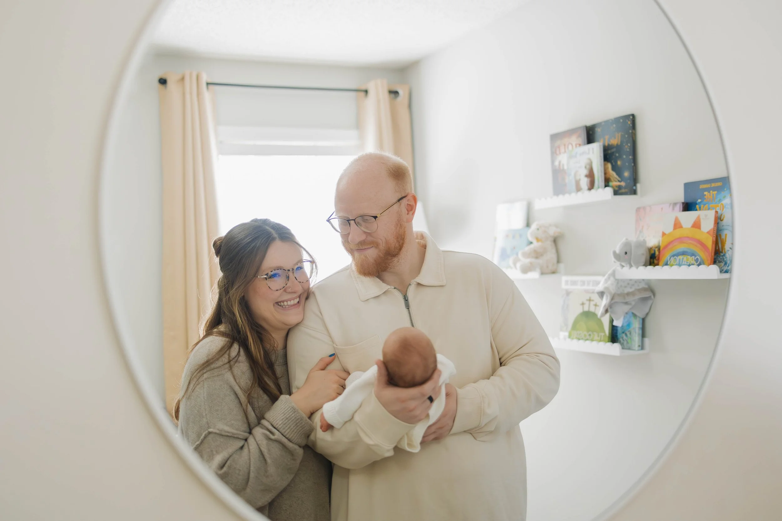 A couple holding a newborn baby is seen through a circular mirror in a room with books and toys on the shelf.