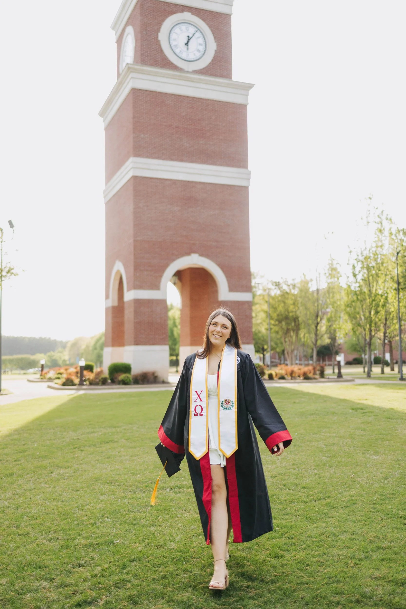 A young woman in a graduation cap and gown walking on a grassy field with a clock tower in the background.
