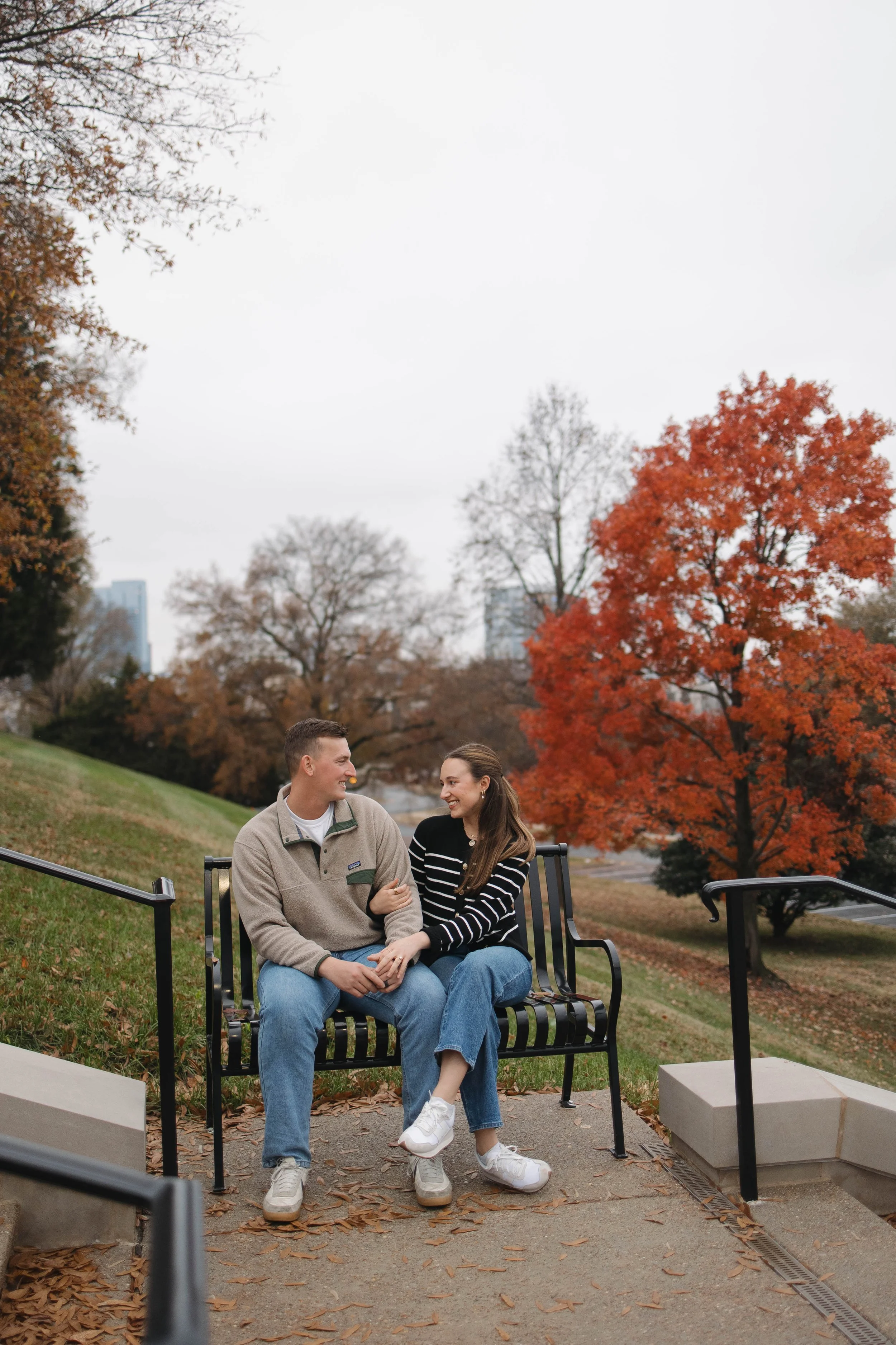 A young man and woman sit on a park bench, holding hands and smiling at each other. The woman is wearing a striped sweater and white sneakers, while the man is dressed in a beige jacket and blue jeans. They are surrounded by autumn trees with colorfu