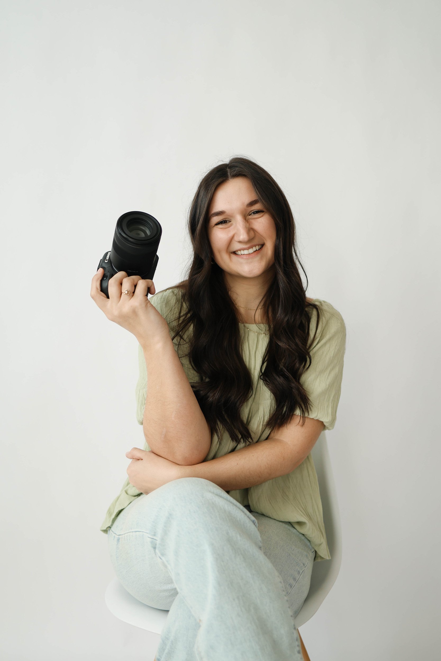 A smiling woman with long dark hair, wearing a light green t-shirt and light-colored jeans, seated in a white chair against a plain white background, holding a camera in her right hand.