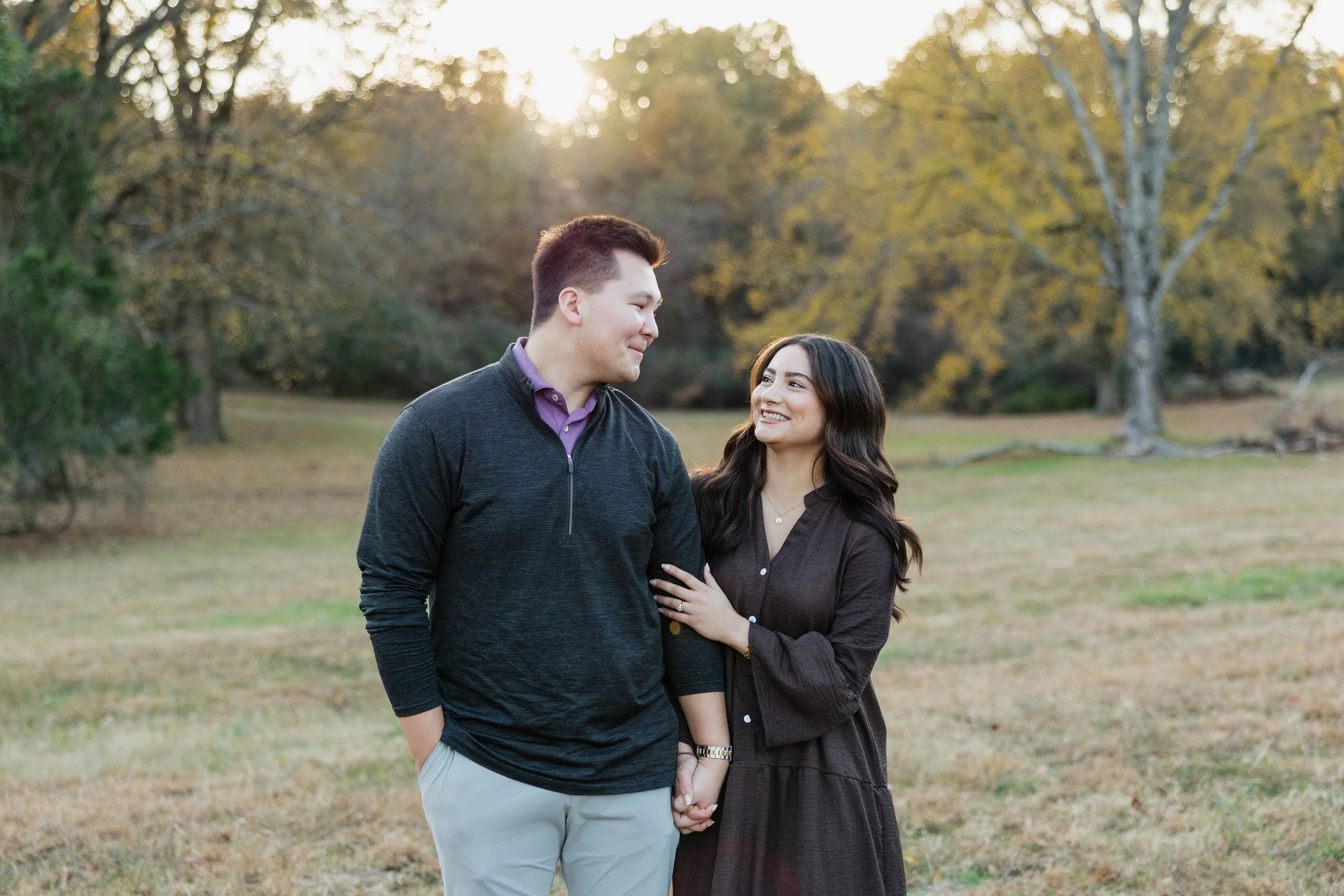 A young couple stands close in a park during autumn, holding hands and smiling at each other, with trees in the background and sunlight filtering through the branches.