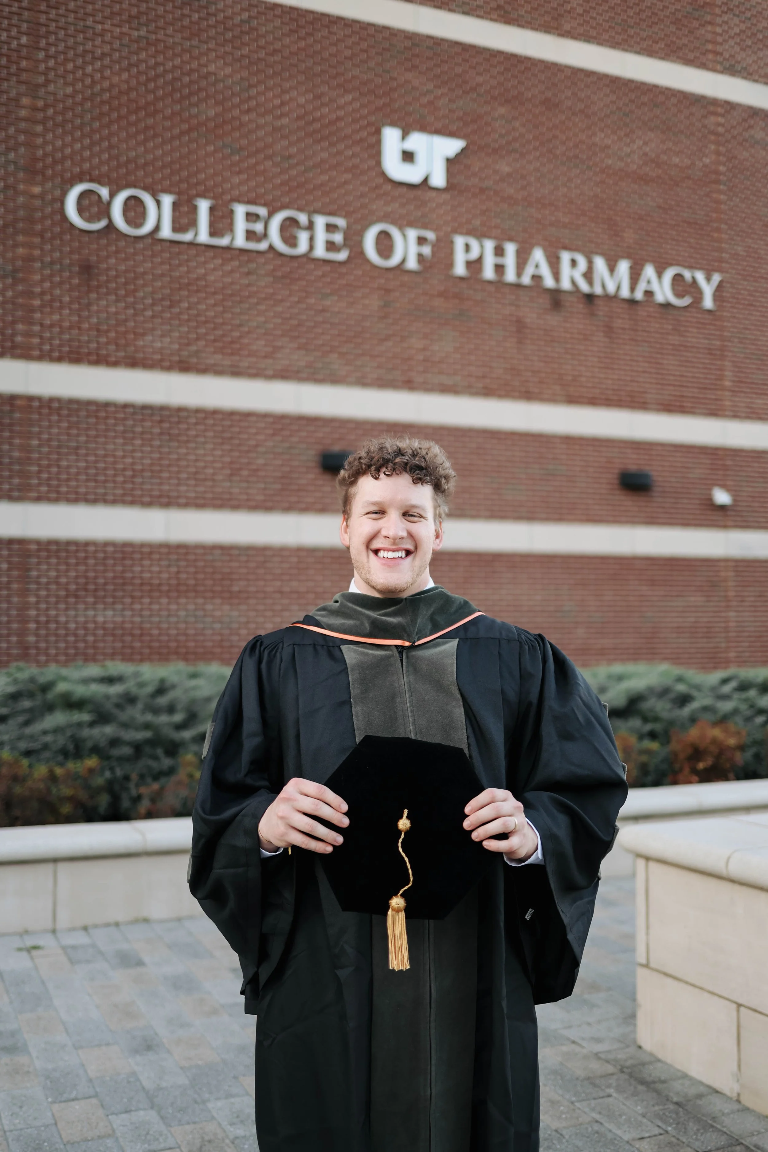A smiling young man in a black graduation gown holding a graduation cap with a gold tassel, standing outdoors in front of a brick building with a sign that reads 'College of Pharmacy'.