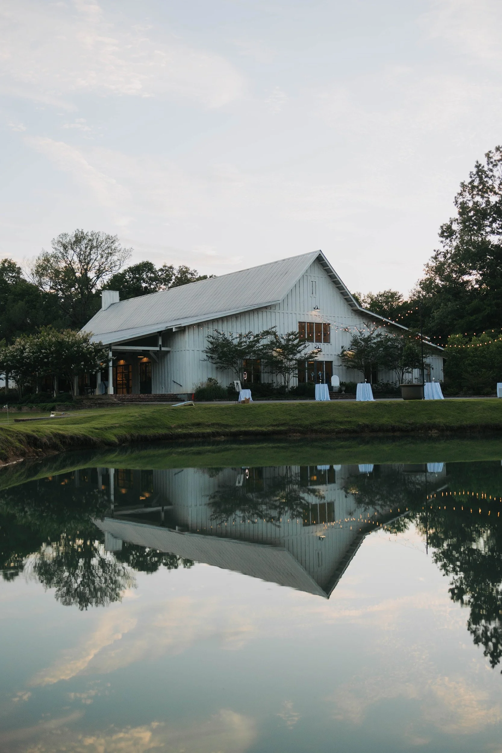 A white barn with string lights, surrounded by trees, and reflected in a body of water during dusk.