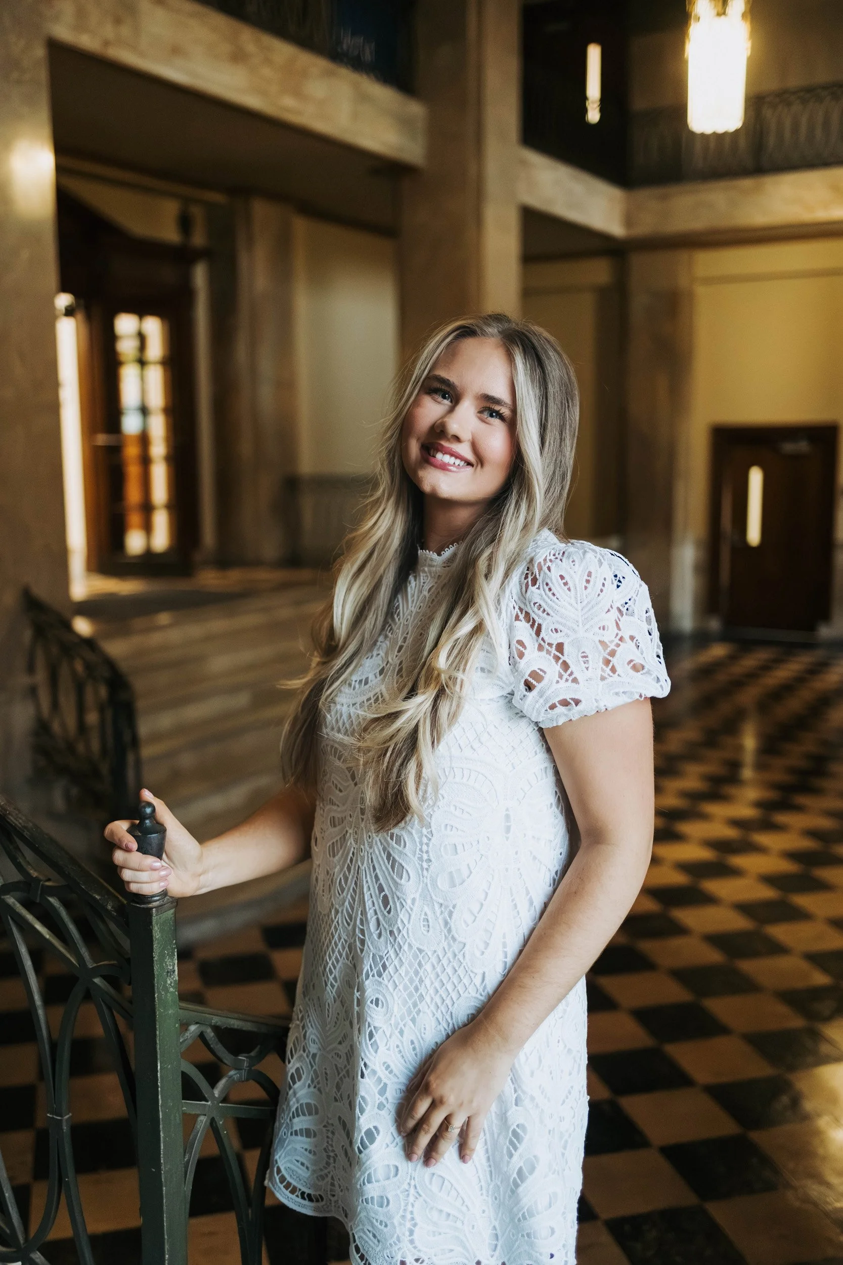 A young woman with long blonde hair, wearing a white lace dress, smiling, standing indoors near a decorative railing.