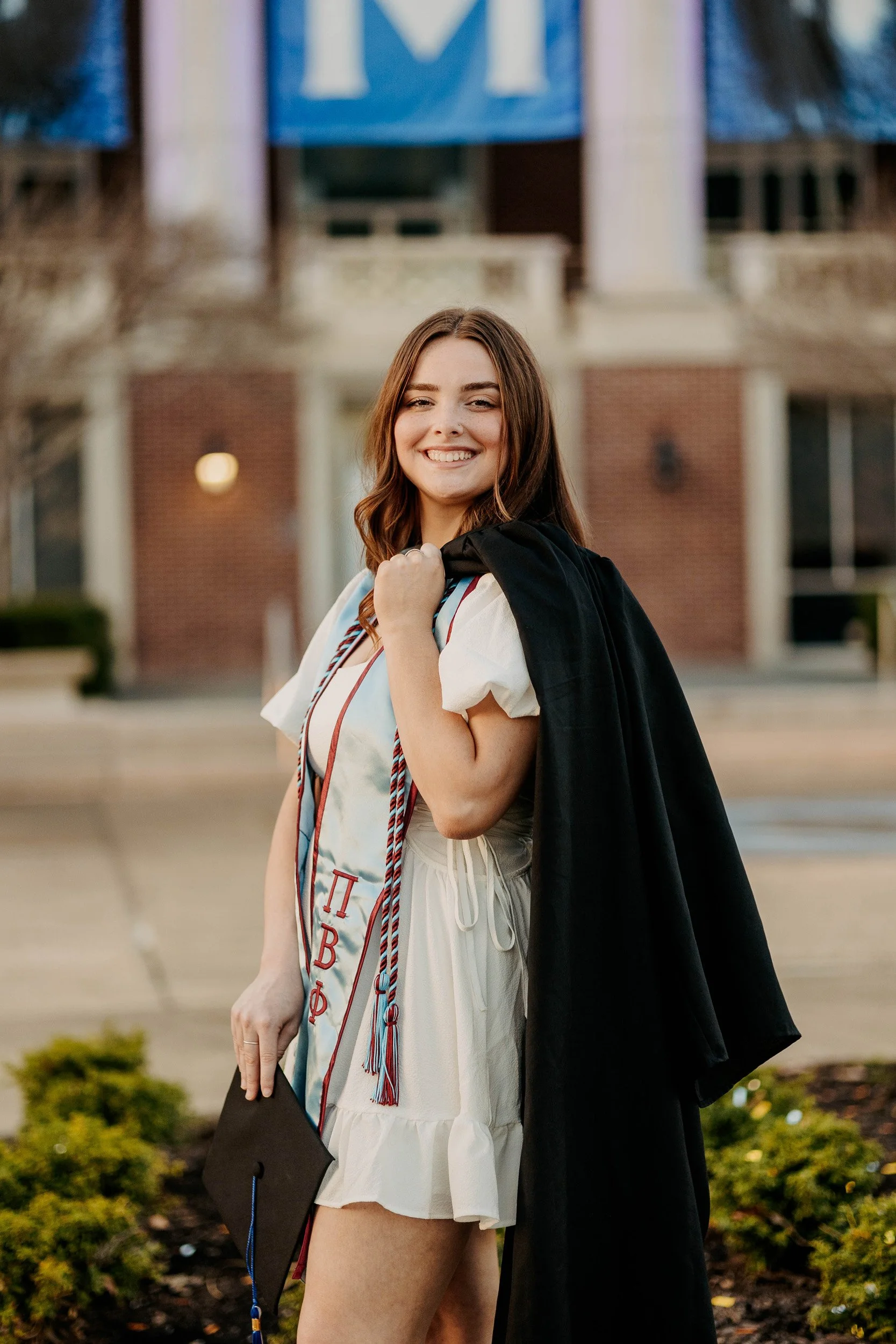 A young woman in a graduation gown and hood smiling outside in front of a brick building with columns, holding a diploma in her left hand and a graduation cap in her right hand.