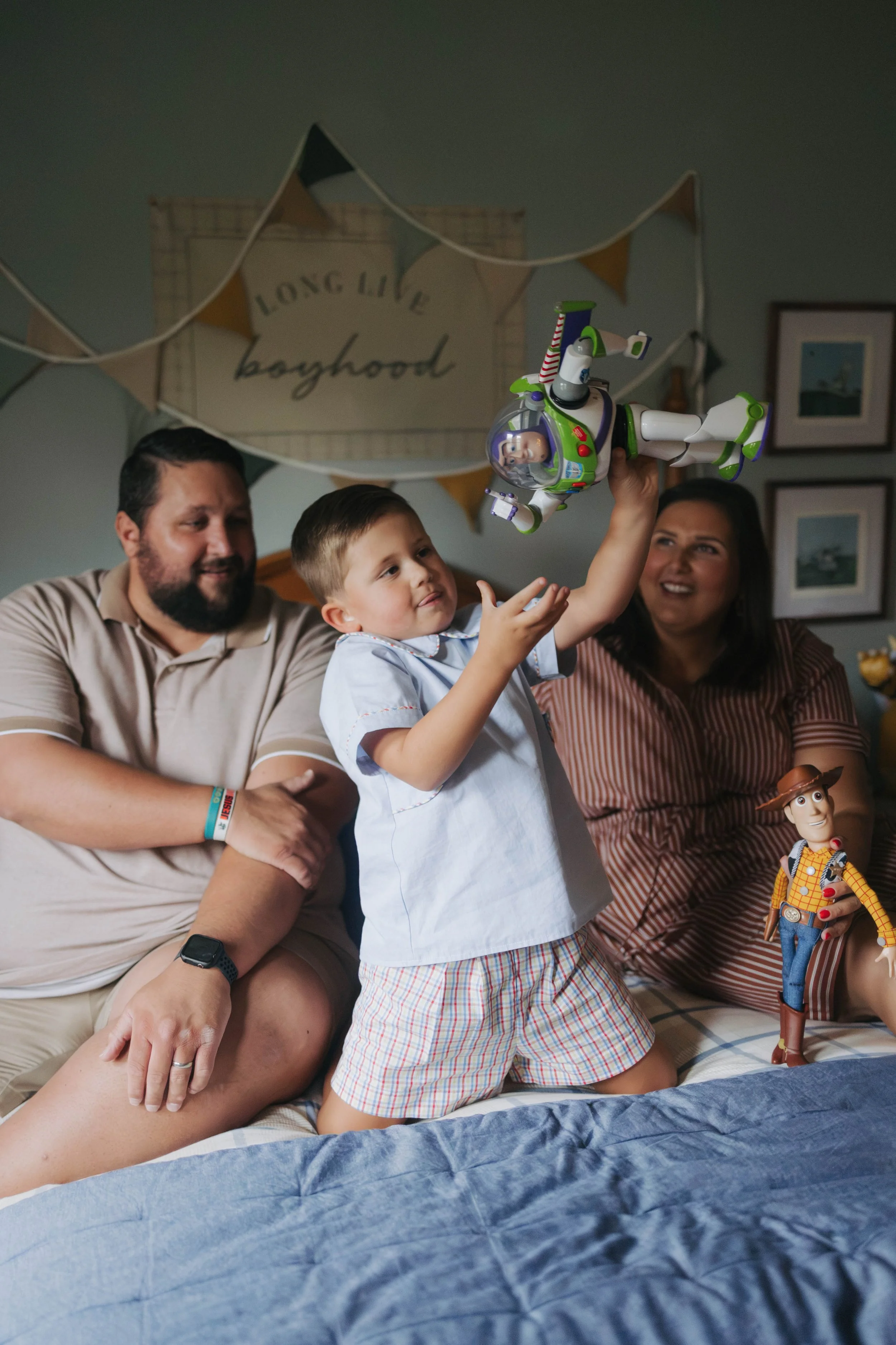 A family of three kids and two adults sitting on a bed playing with character toys, including Buzz Lightyear from Toy Story, in a decorated room with a banner saying 'Long Live Boyhood'.