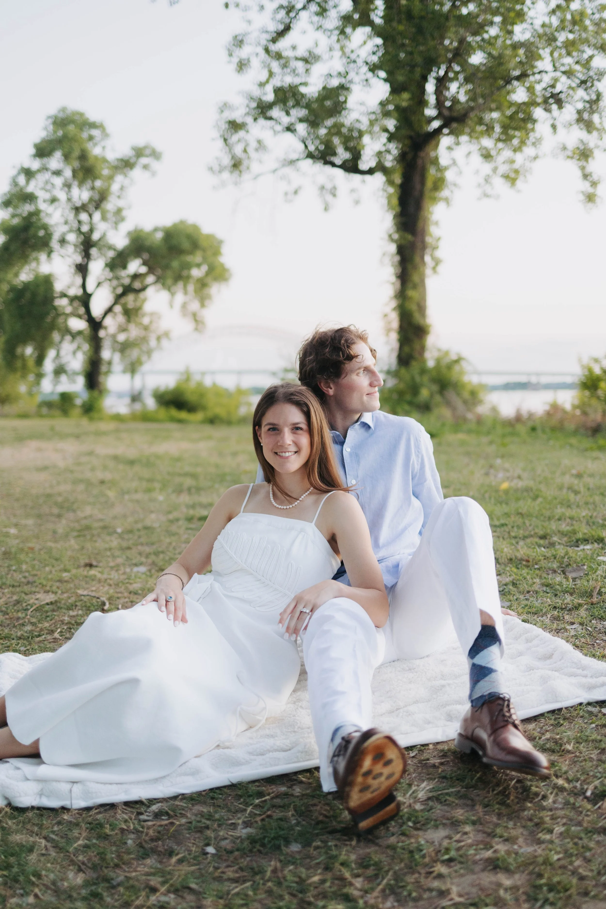 A young couple, dressed in wedding attire, sitting on a white blanket in a park with lush green trees, smiling and enjoying a moment together.