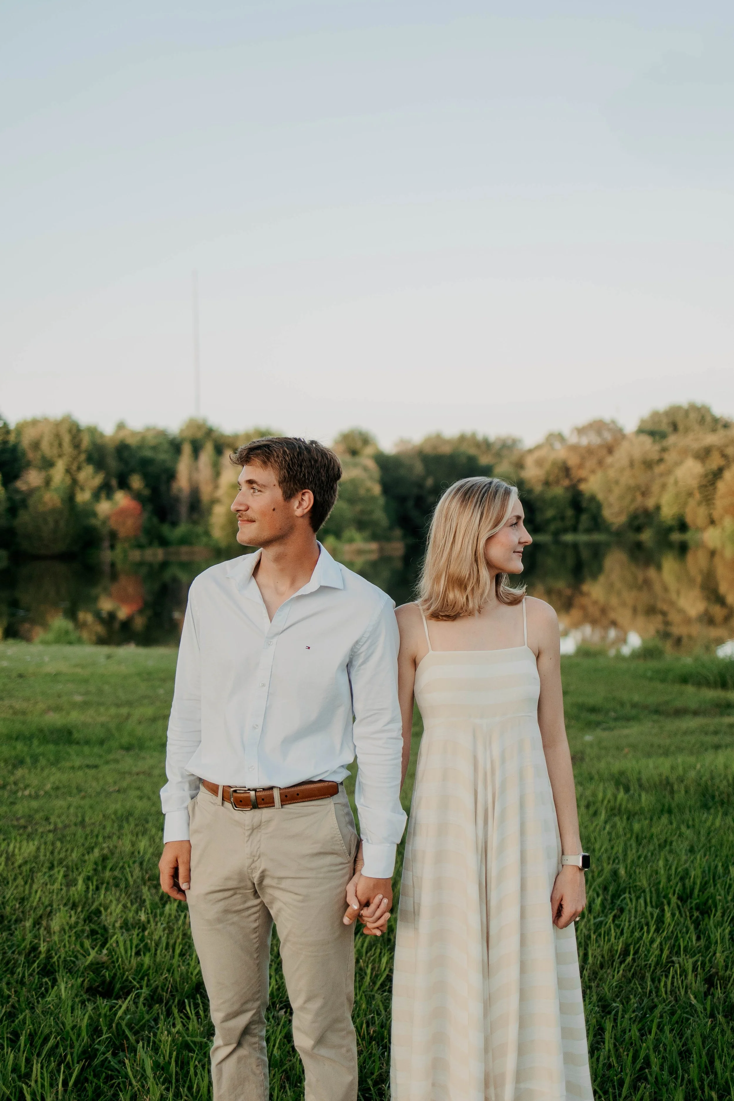 A young man and woman are standing outdoors on grass near a body of water, holding hands and looking in opposite directions, with trees and a clear sky in the background.