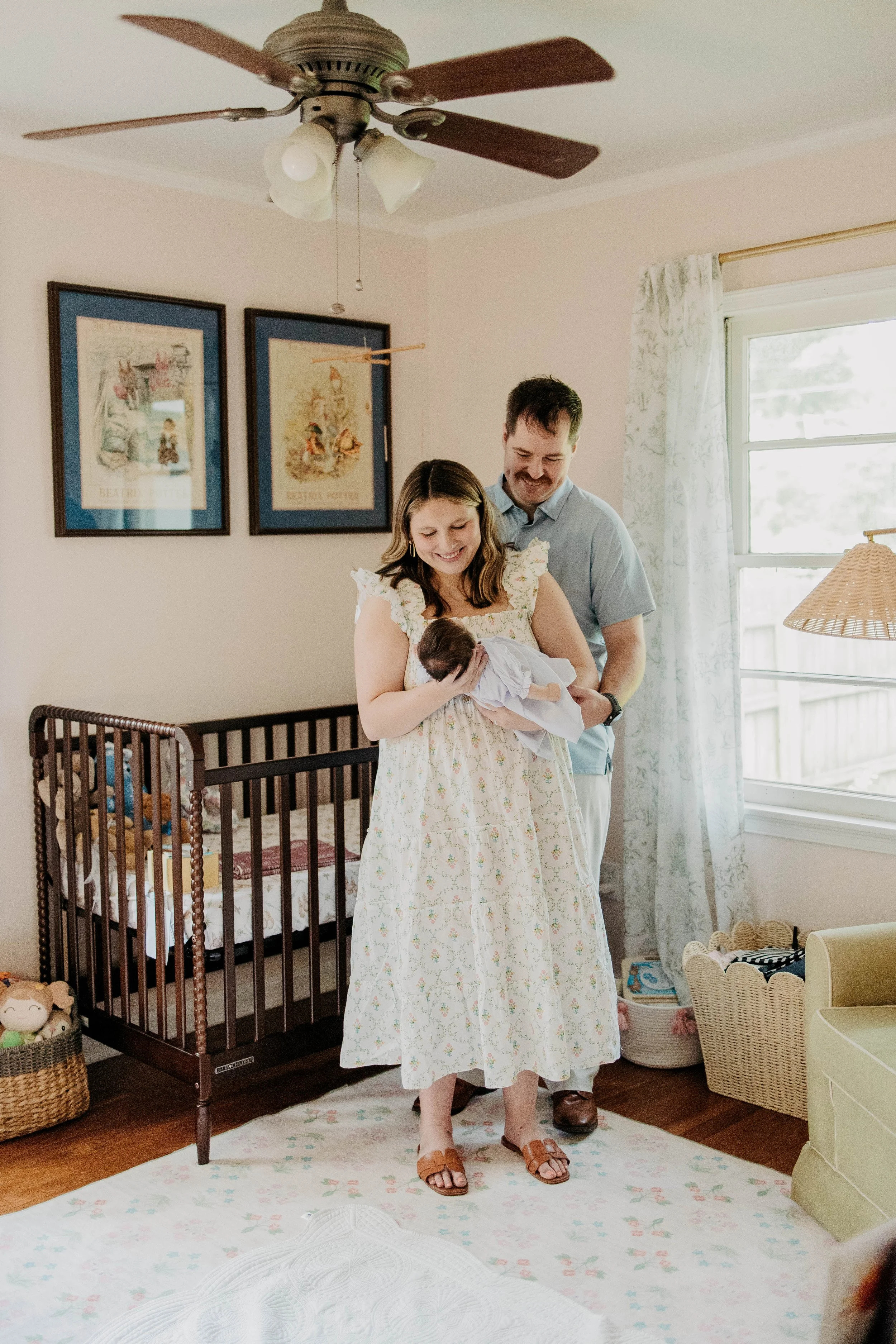 A woman and a man are standing in a nursery, holding a newborn baby. The woman is smiling at the baby, wearing a floral dress and sandals. The man is smiling at the baby, standing behind her, wearing a light blue shirt and brown shoes. There is a cri