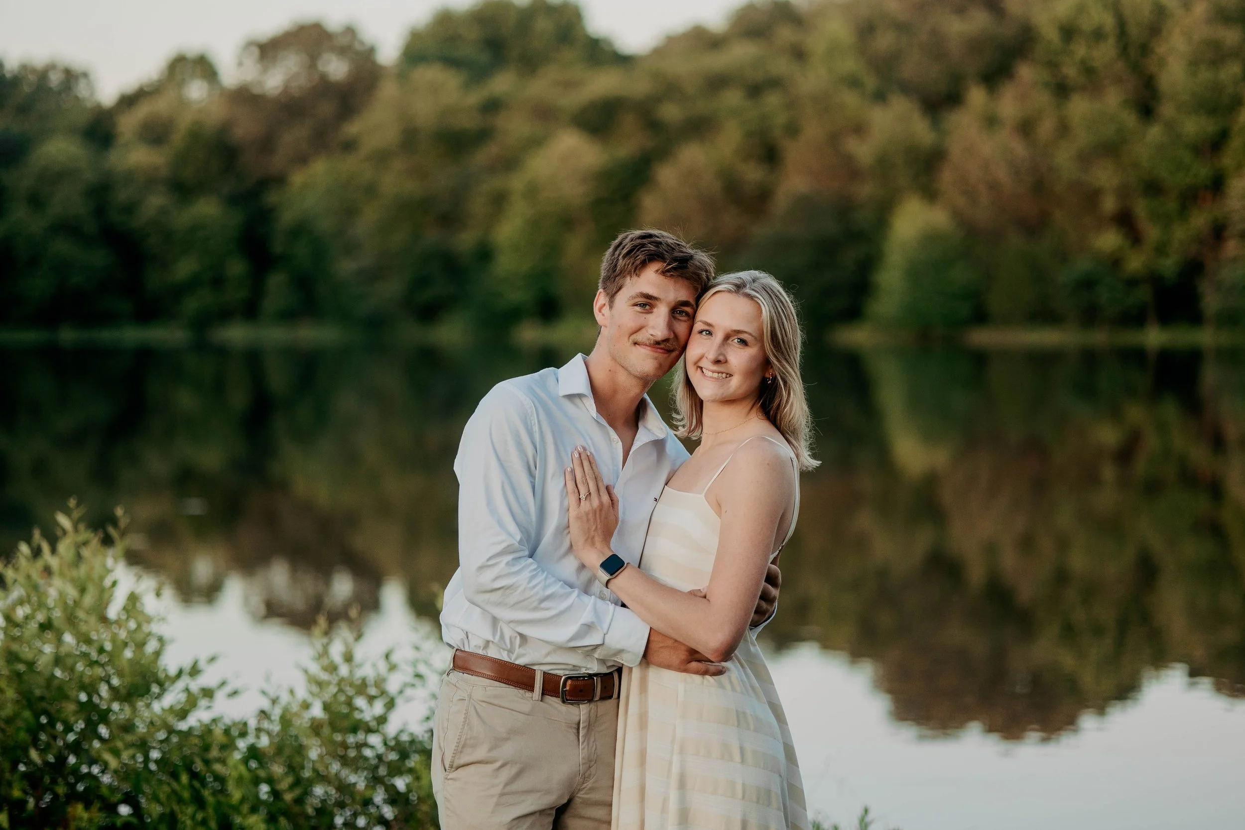 A happy couple embracing outdoors near a lake with trees in the background, during the daytime.