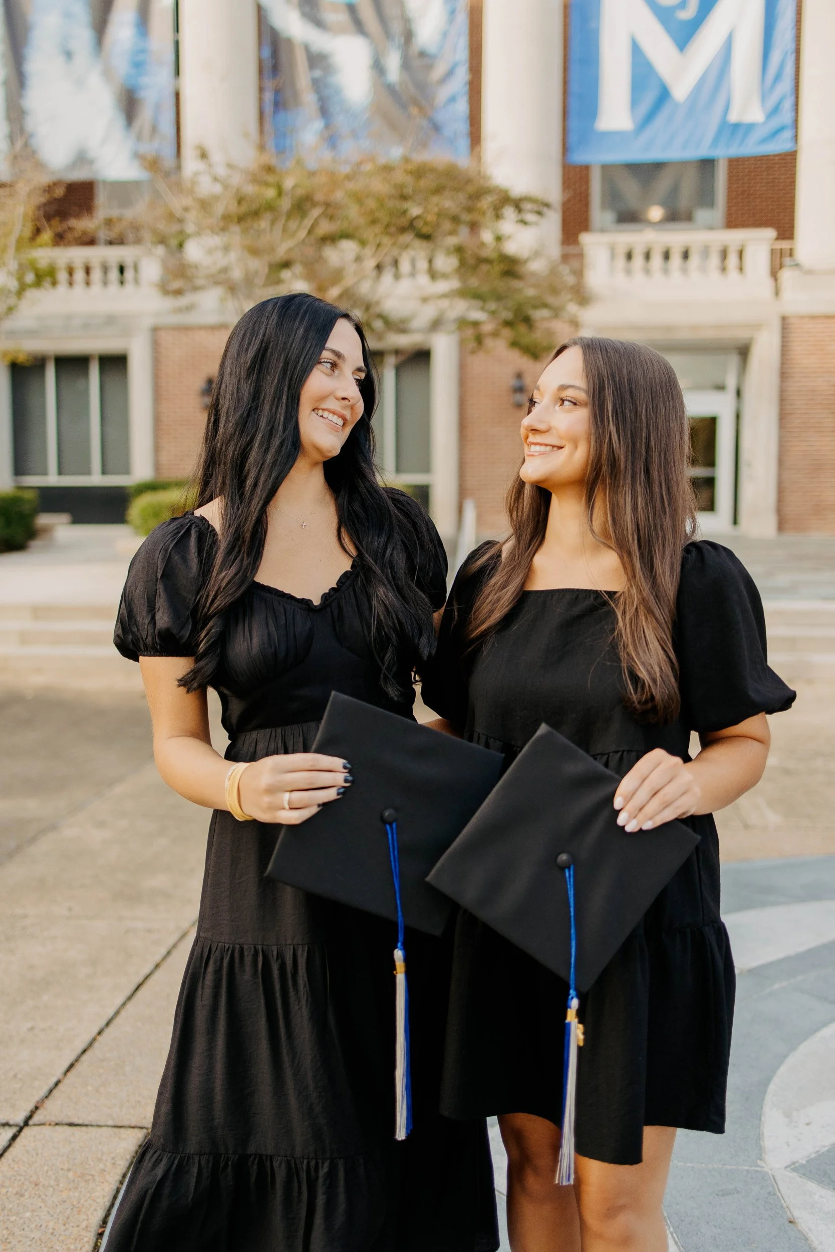 Two young women in black graduation dresses holding caps and diplomas stand outside in front of a building with large windows and a blue banner that says 'M'. They are smiling at each other.