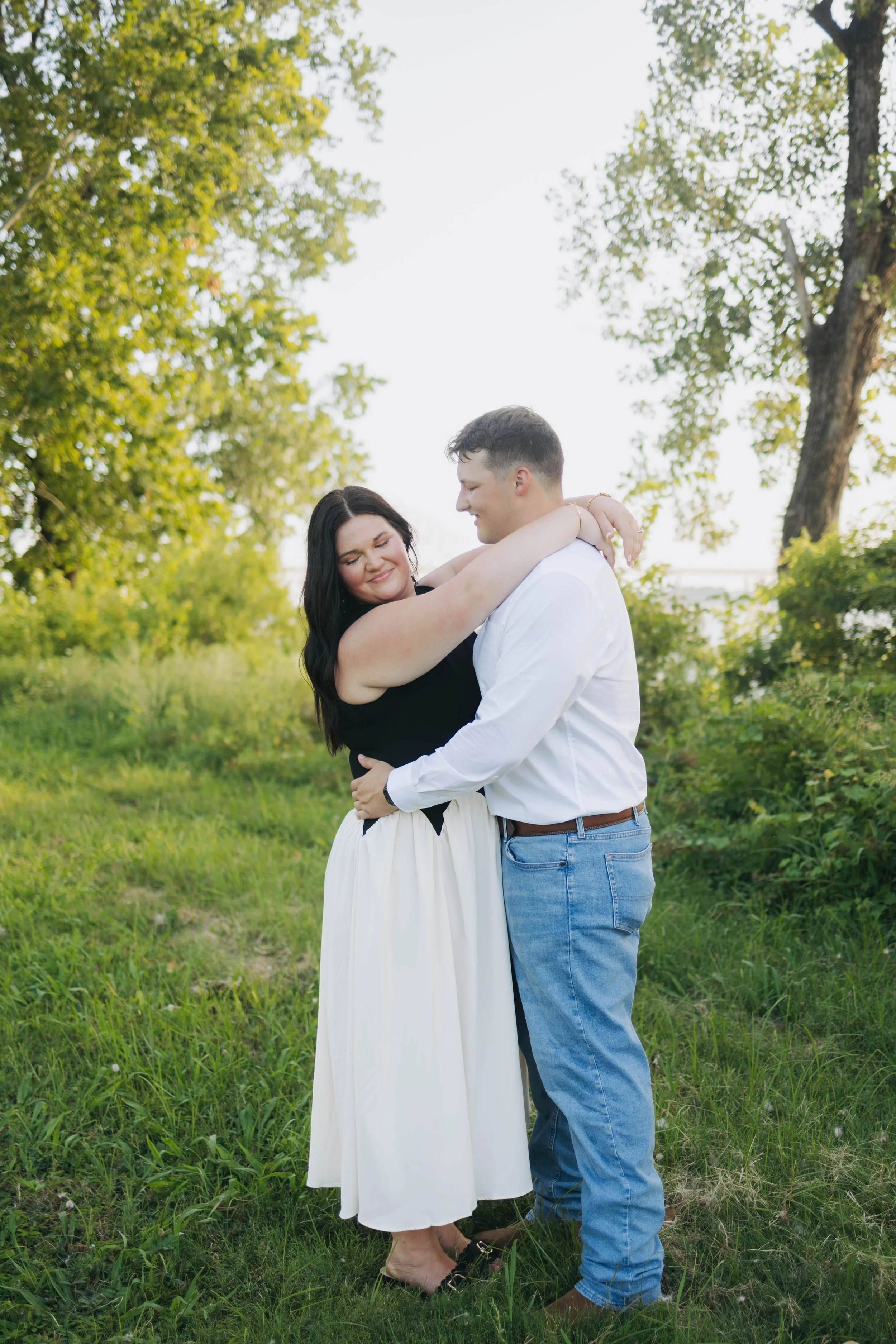 A couple embracing each other outdoors in a grassy area with trees, smiling and looking at each other.