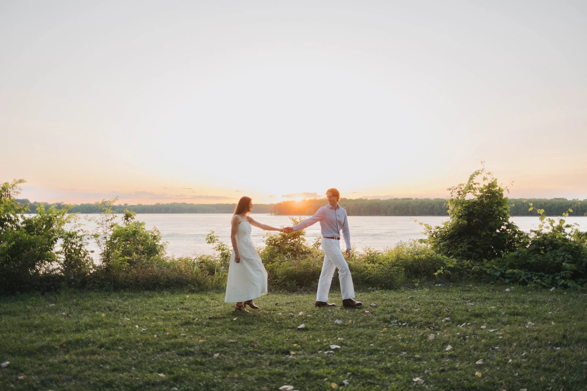 A couple dressed in wedding attire walking hand-in-hand along a grassy area near a body of water during sunset.