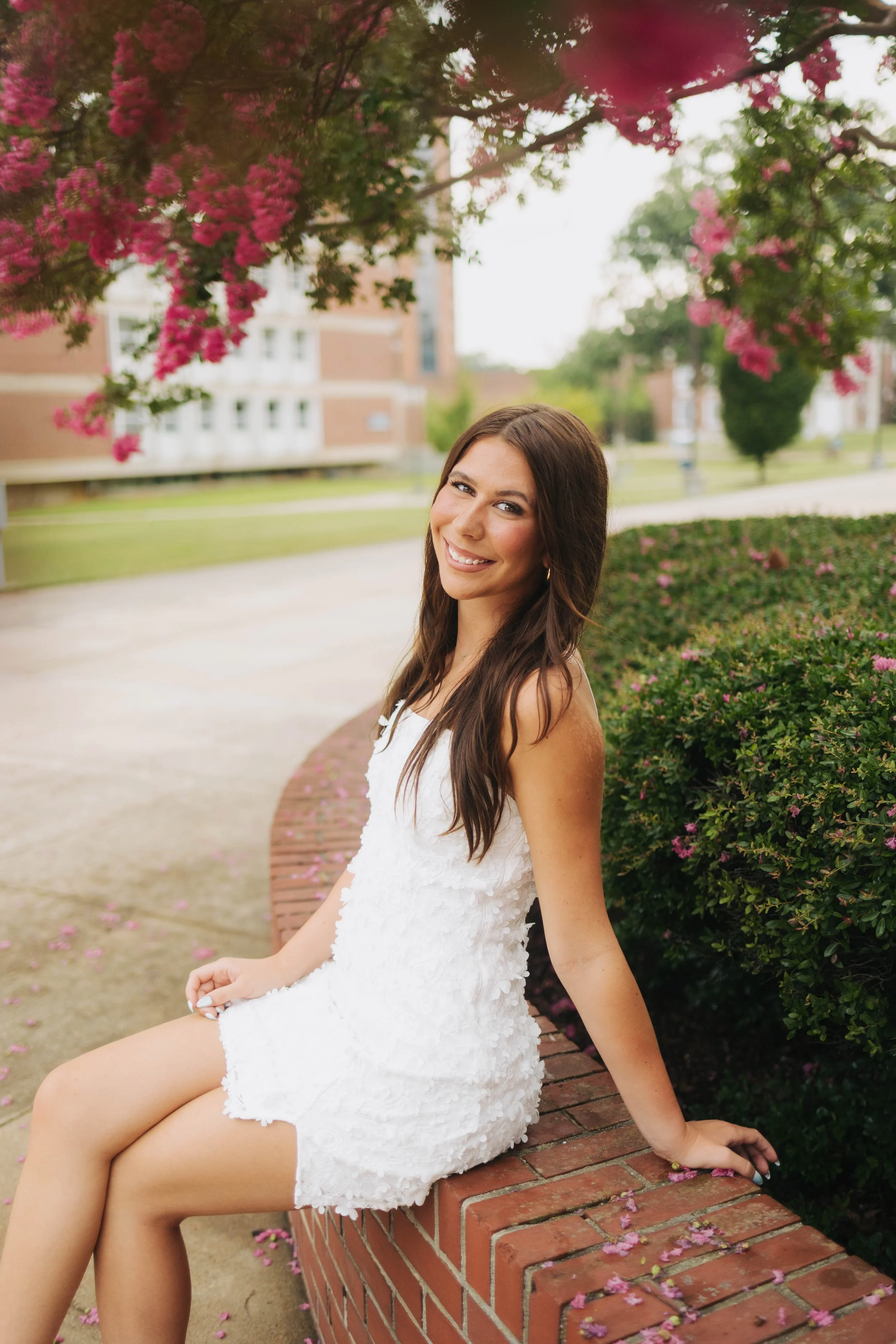 A young woman with long brown hair sitting on a brick wall in a white lace dress, smiling, with pink flowers on trees above and scattered on the ground, in a park-like setting.