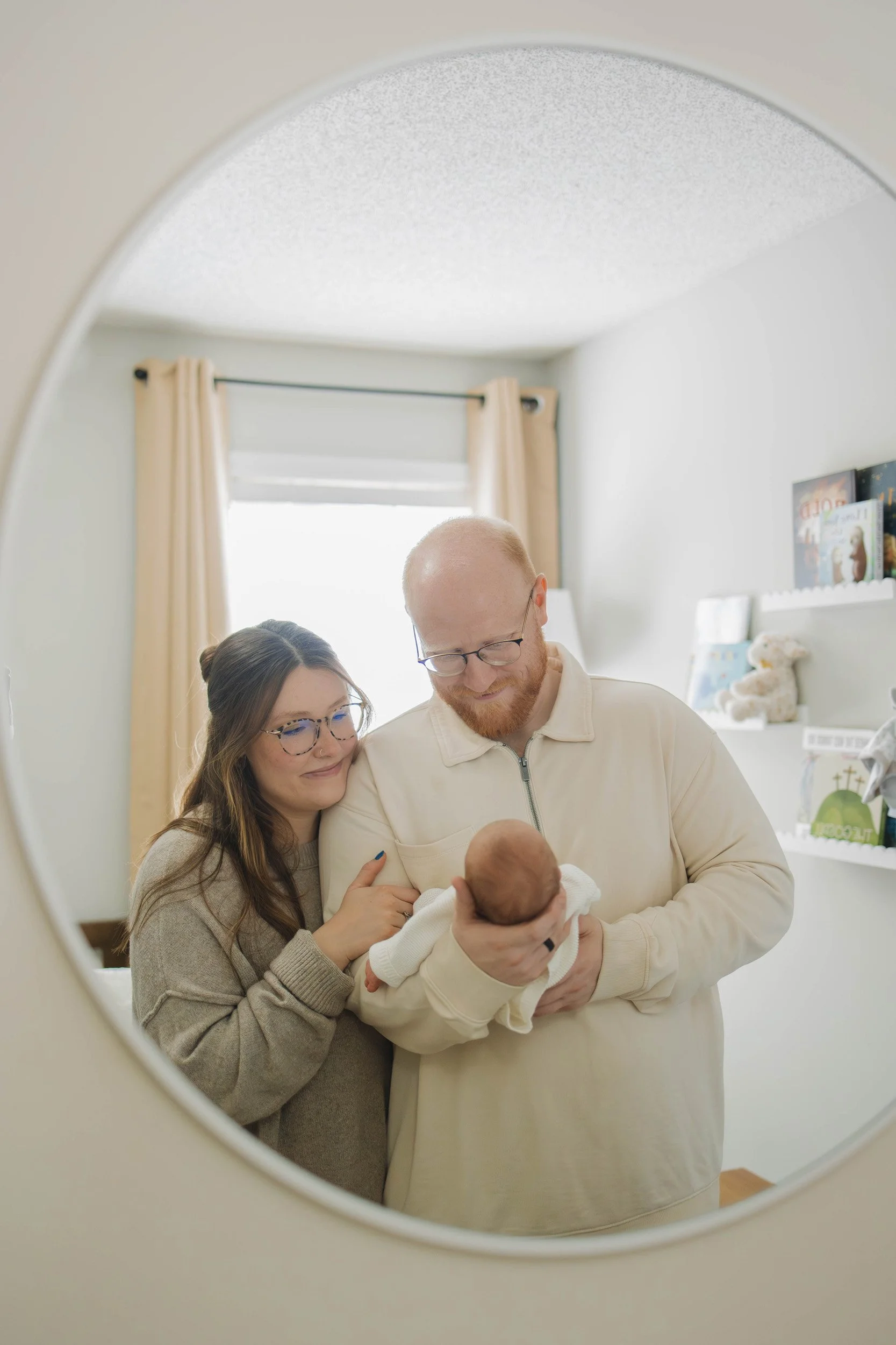 A couple is holding their newborn baby during a gentle moment, as seen through a round mirror in a cozy, well-lit room with cream-colored curtains and children's books.