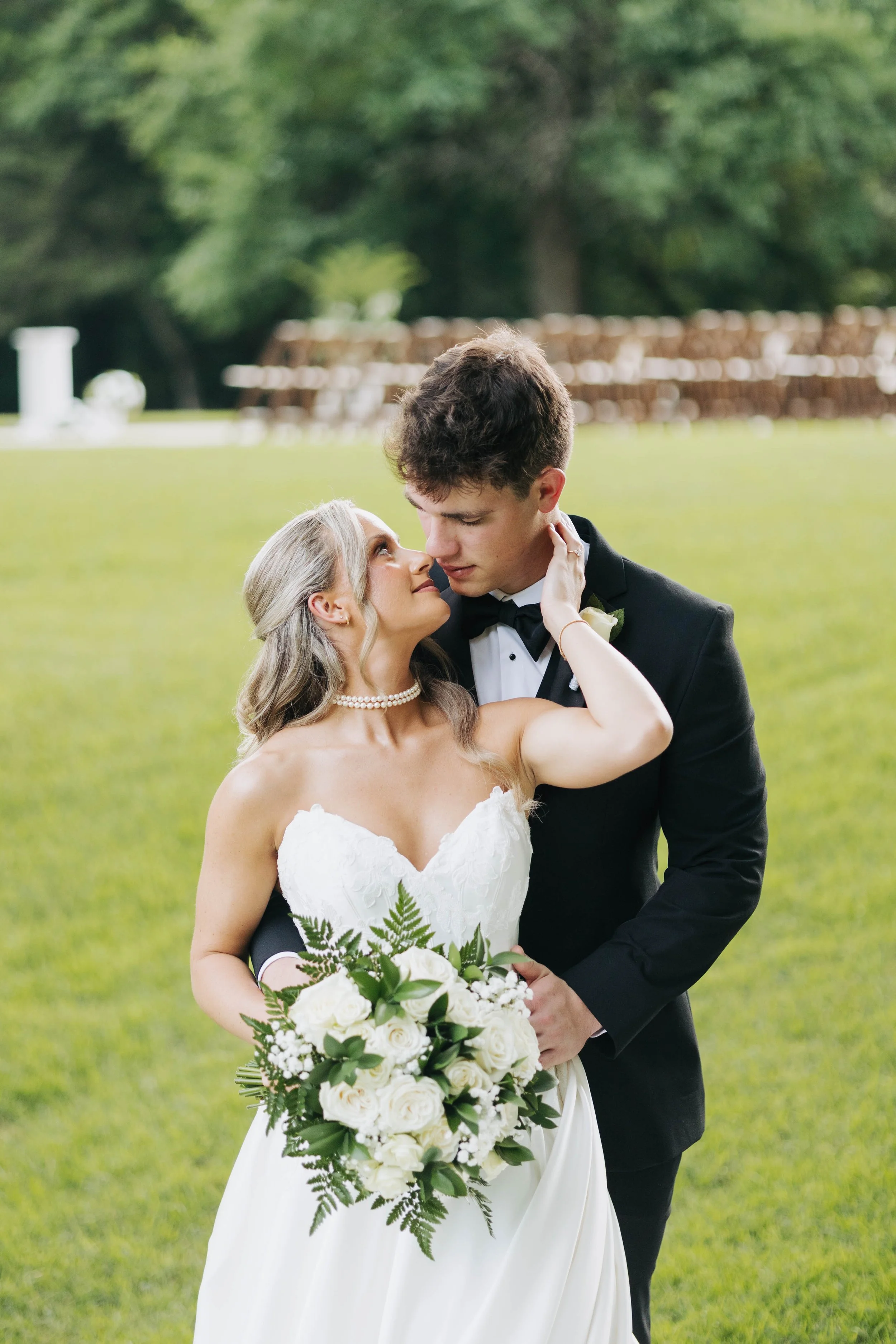 A bride and groom share an intimate moment outdoors on their wedding day, with the bride holding a large bouquet of white roses and greenery, and the groom dressed in a black tuxedo leaning in towards her.